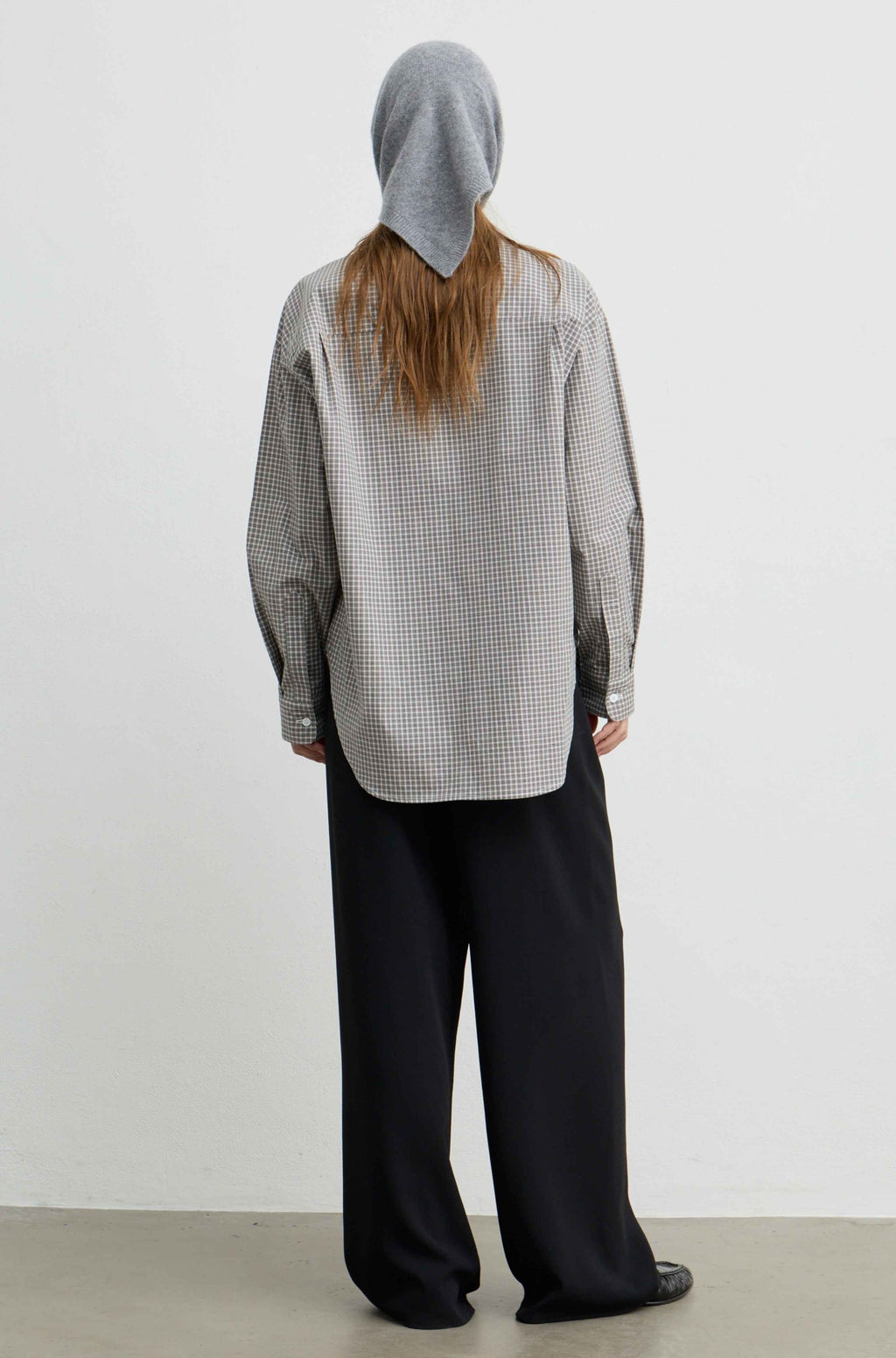 A model shown from the back wearing a light gray and black gingham patterned long-sleeve shirt with a gray knit hood, paired with loose black trousers, against a plain white studio background.