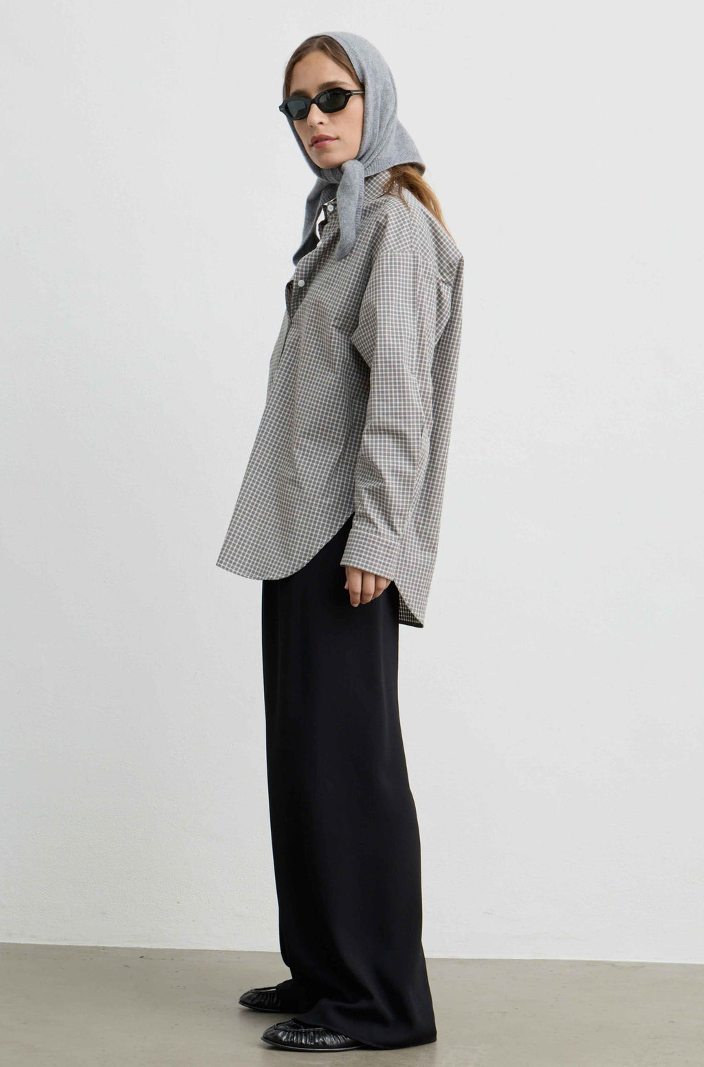 A woman wearing a gray and white gingham patterned button-up shirt, black wide-leg trousers, a light gray hijab, and black sunglasses, standing in profile against a plain light gray studio background.
