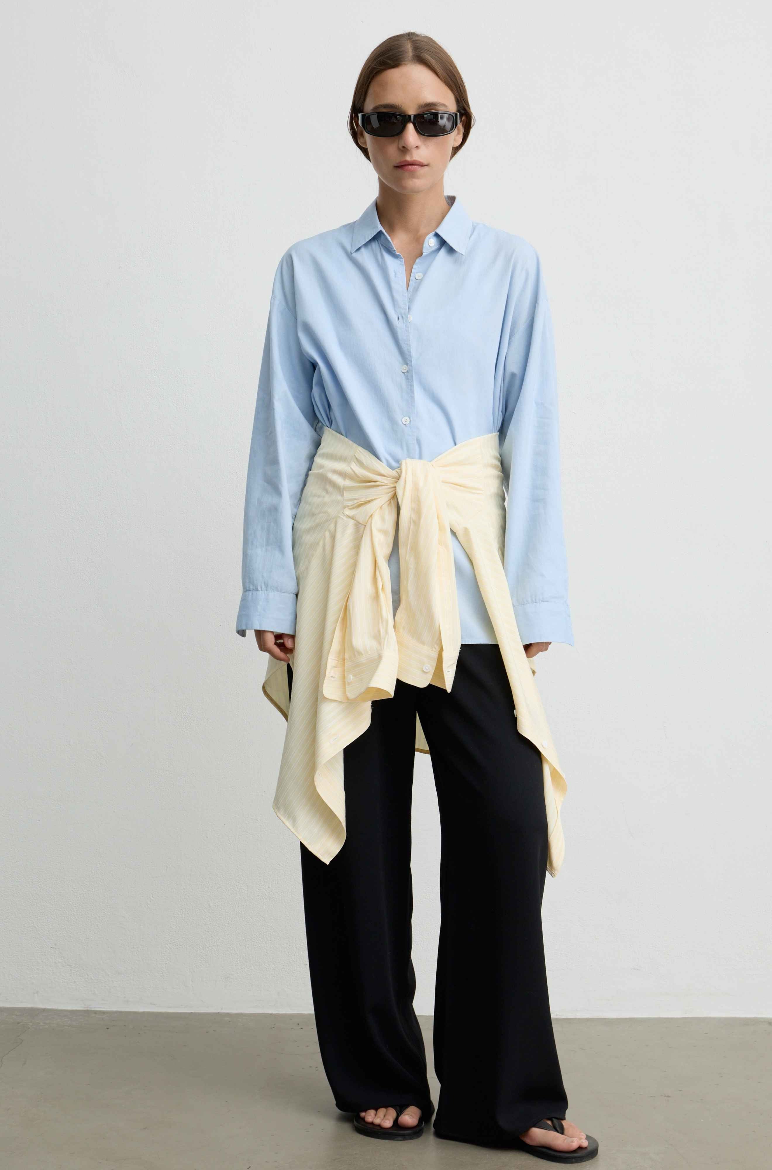 A model stands against a plain light gray studio wall wearing a light blue button-up shirt, black wide-leg pants, and black sandals, with a cream wrap tied around the waist.