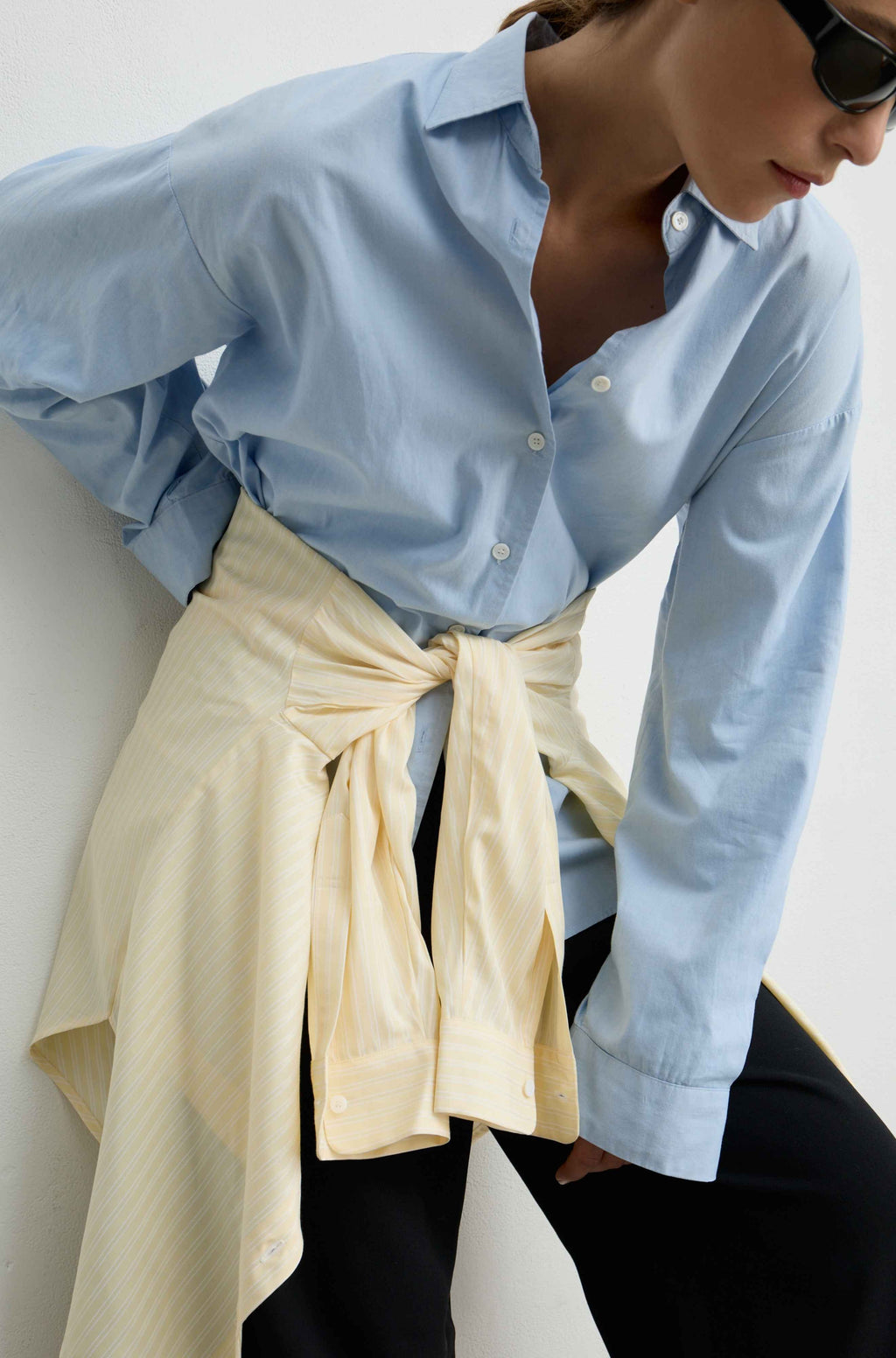 A woman wearing a light blue long-sleeve button-up shirt tucked into black pants, with a cream-colored striped shirt tied around her waist, photographed against a plain light background.