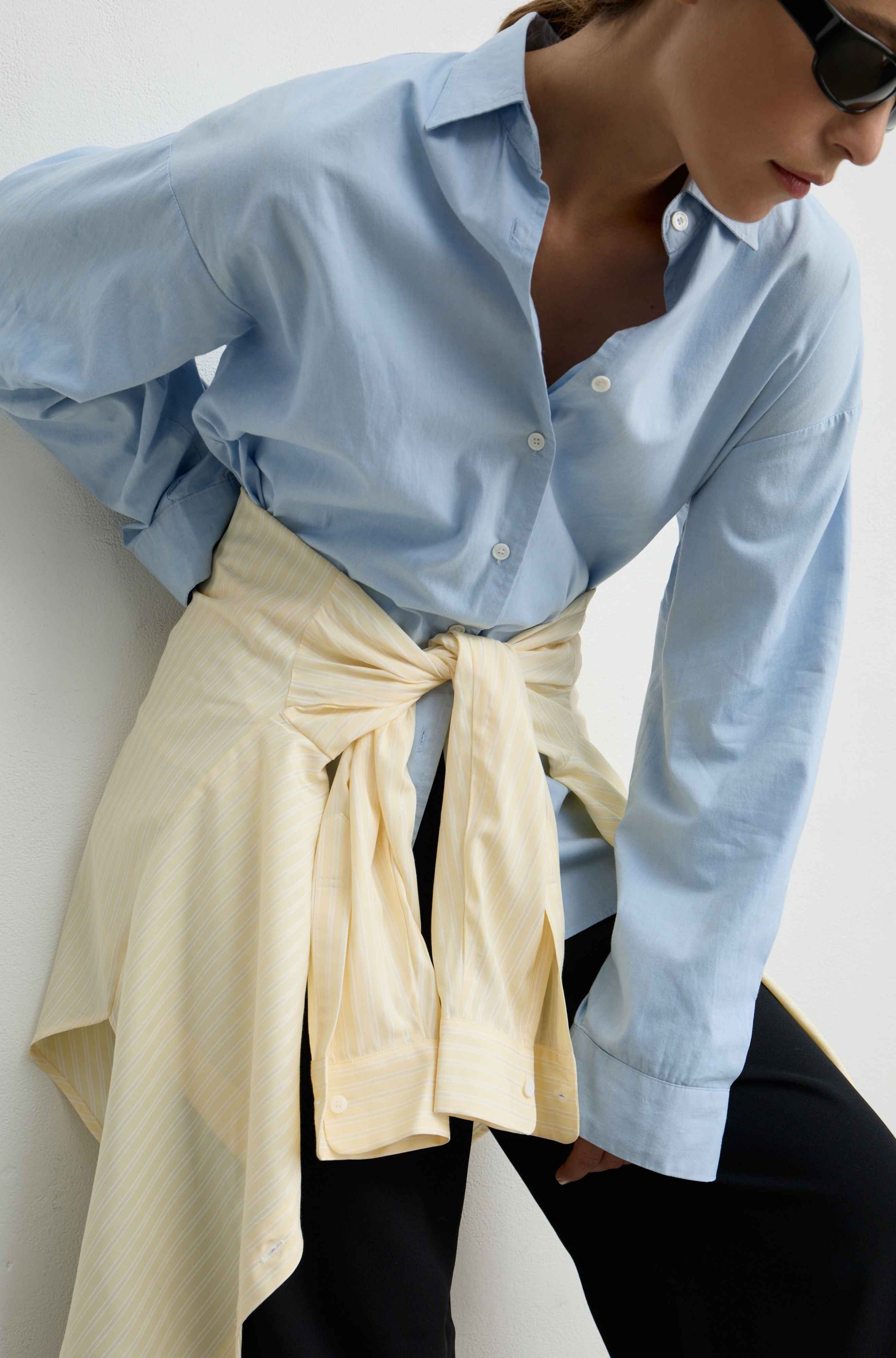 A woman wearing a light blue long-sleeve button-up shirt tucked into black pants, with a cream-colored striped shirt tied around her waist, photographed against a plain light background.