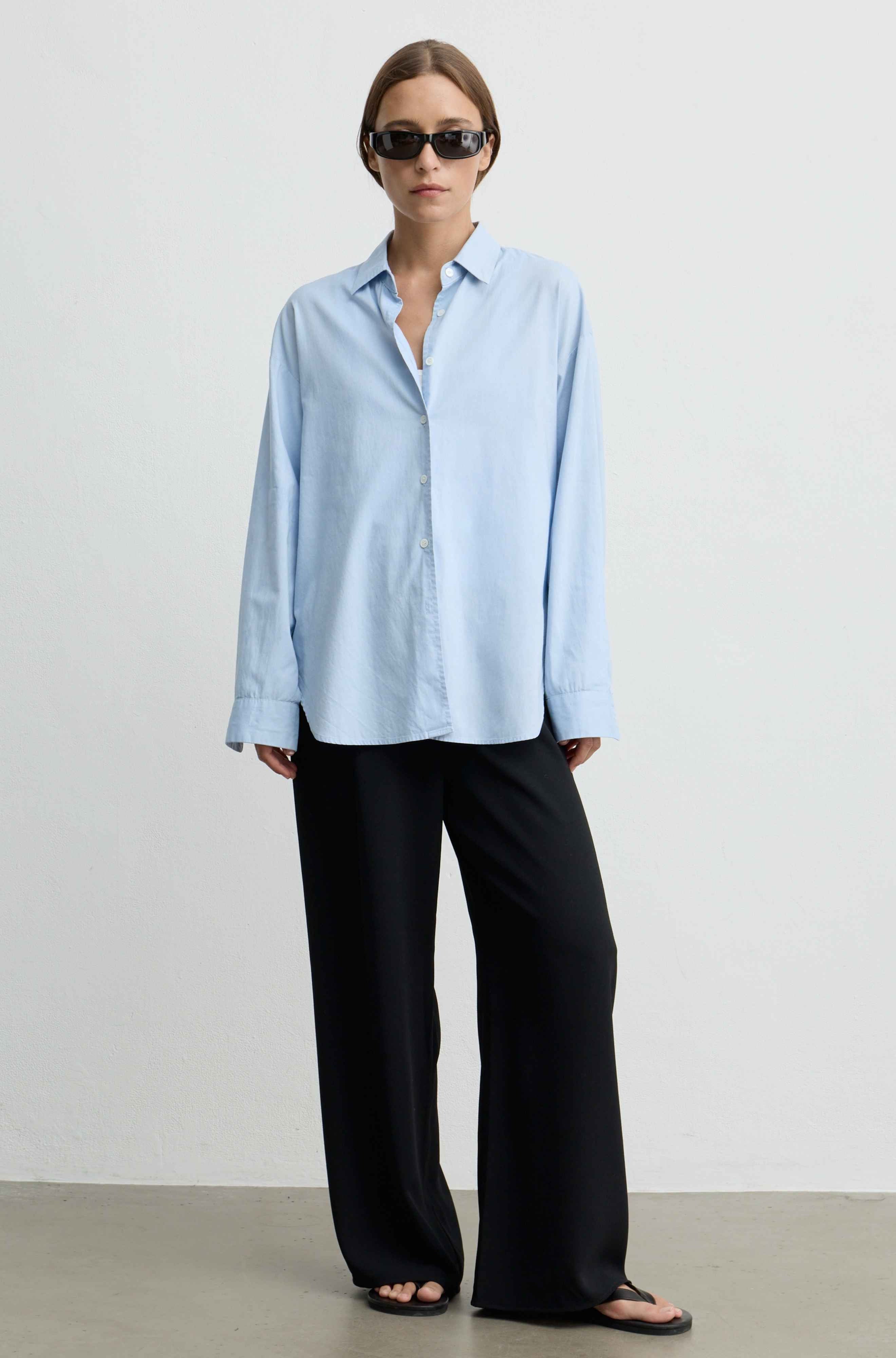 A woman modeling a light blue long-sleeve button-up shirt with black wide-leg trousers and black sandals, standing against a plain light gray studio background.