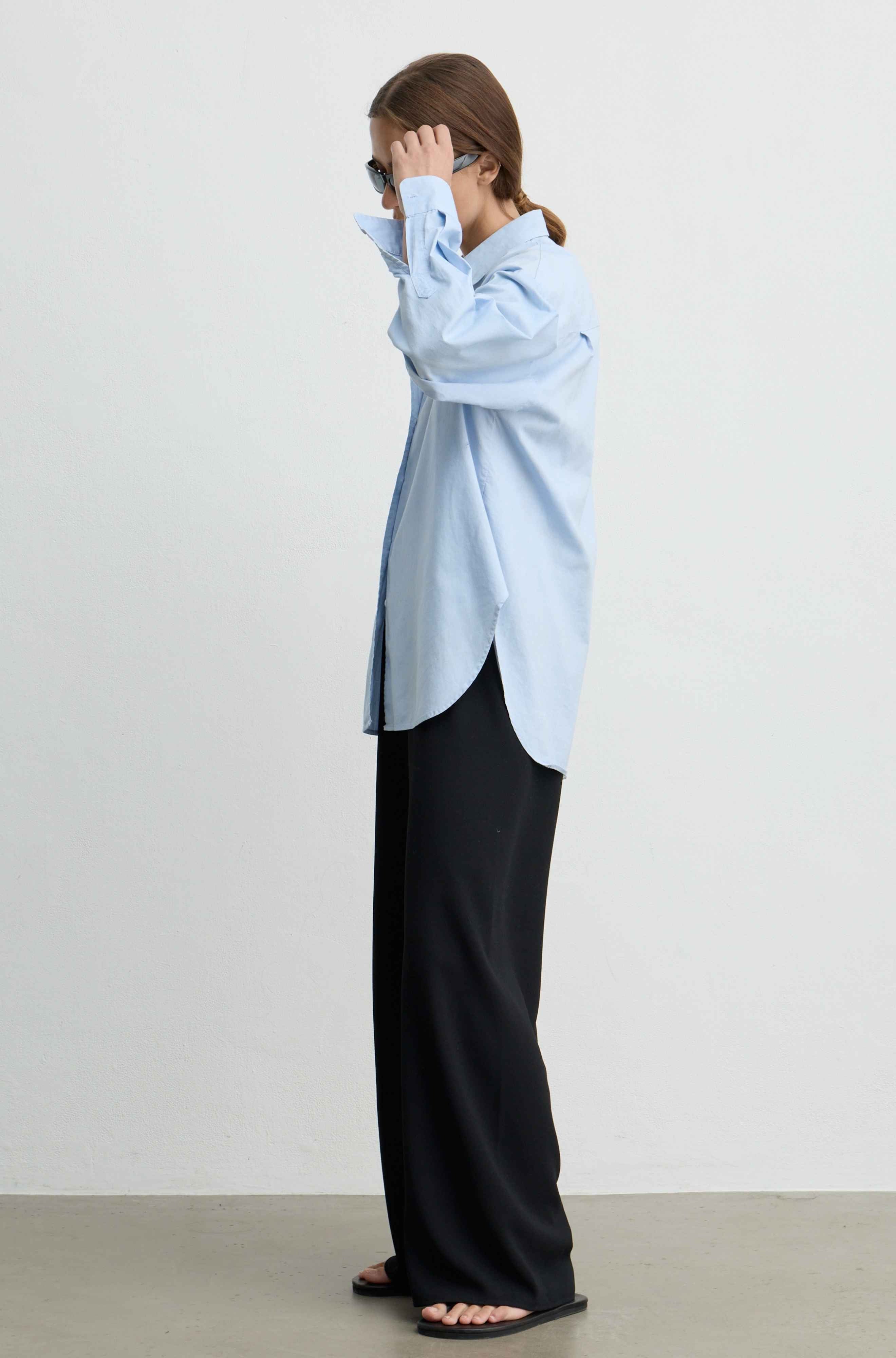 A woman stands in profile wearing an oversized light blue button-up shirt and loose black wide-leg pants against a plain light gray studio background.