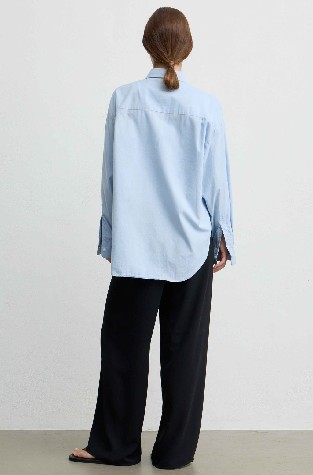 A model shown from the back wearing a light blue button-down shirt and black wide-leg trousers, standing against a plain off-white wall.