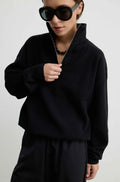 A model wearing a black long-sleeve quarter-zip pullover sweater and matching black trousers, styled with oversized black sunglasses and a black beaded necklace against a plain light gray background.