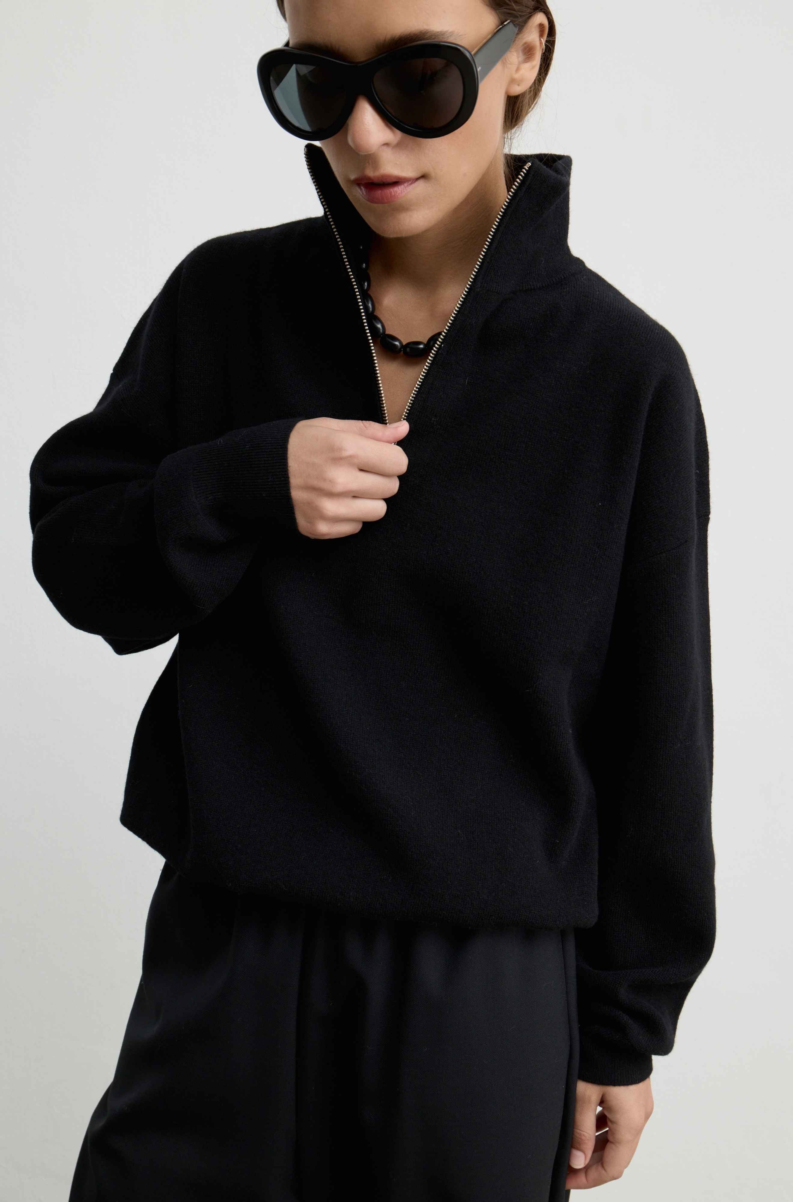 A model wearing a black long-sleeve quarter-zip pullover sweater and matching black trousers, styled with oversized black sunglasses and a black beaded necklace against a plain light gray background.