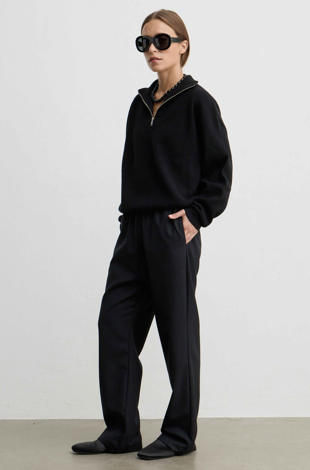 A model wearing a black half-zip sweatshirt and matching black wide-leg sweatpants, styled with black sunglasses and black shoes, standing against a plain light gray studio background.