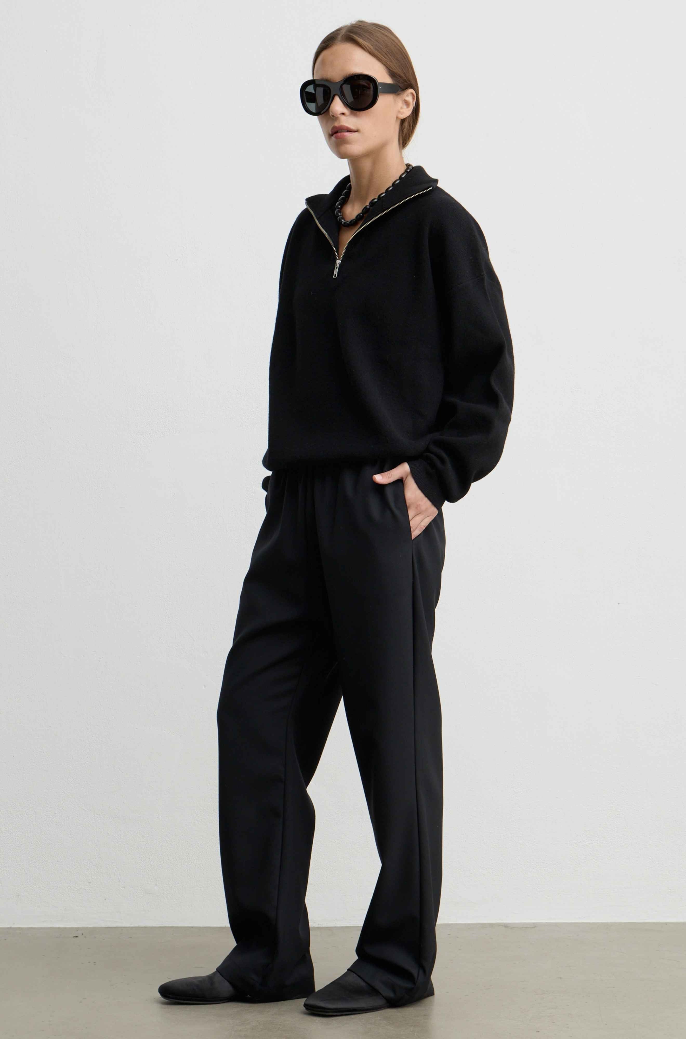 A model wearing a black half-zip sweatshirt and matching black wide-leg sweatpants, styled with black sunglasses and black shoes, standing against a plain light gray studio background.