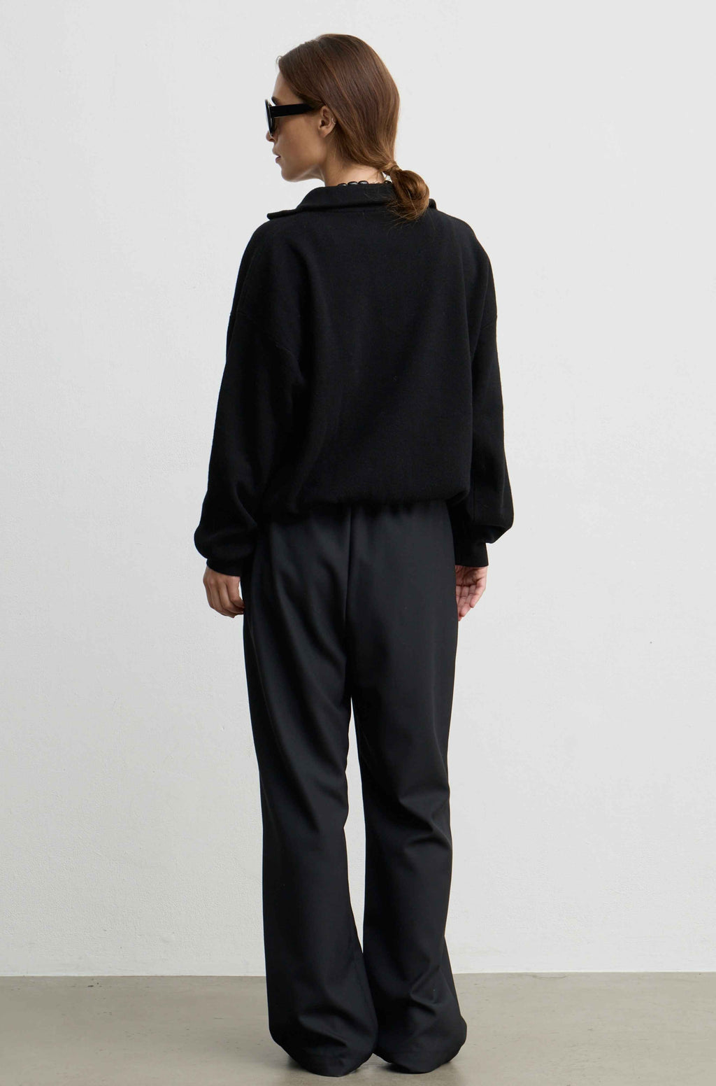 A model viewed from the back wearing a black long-sleeve collared top and matching black wide-leg trousers, standing against a plain light gray studio wall.