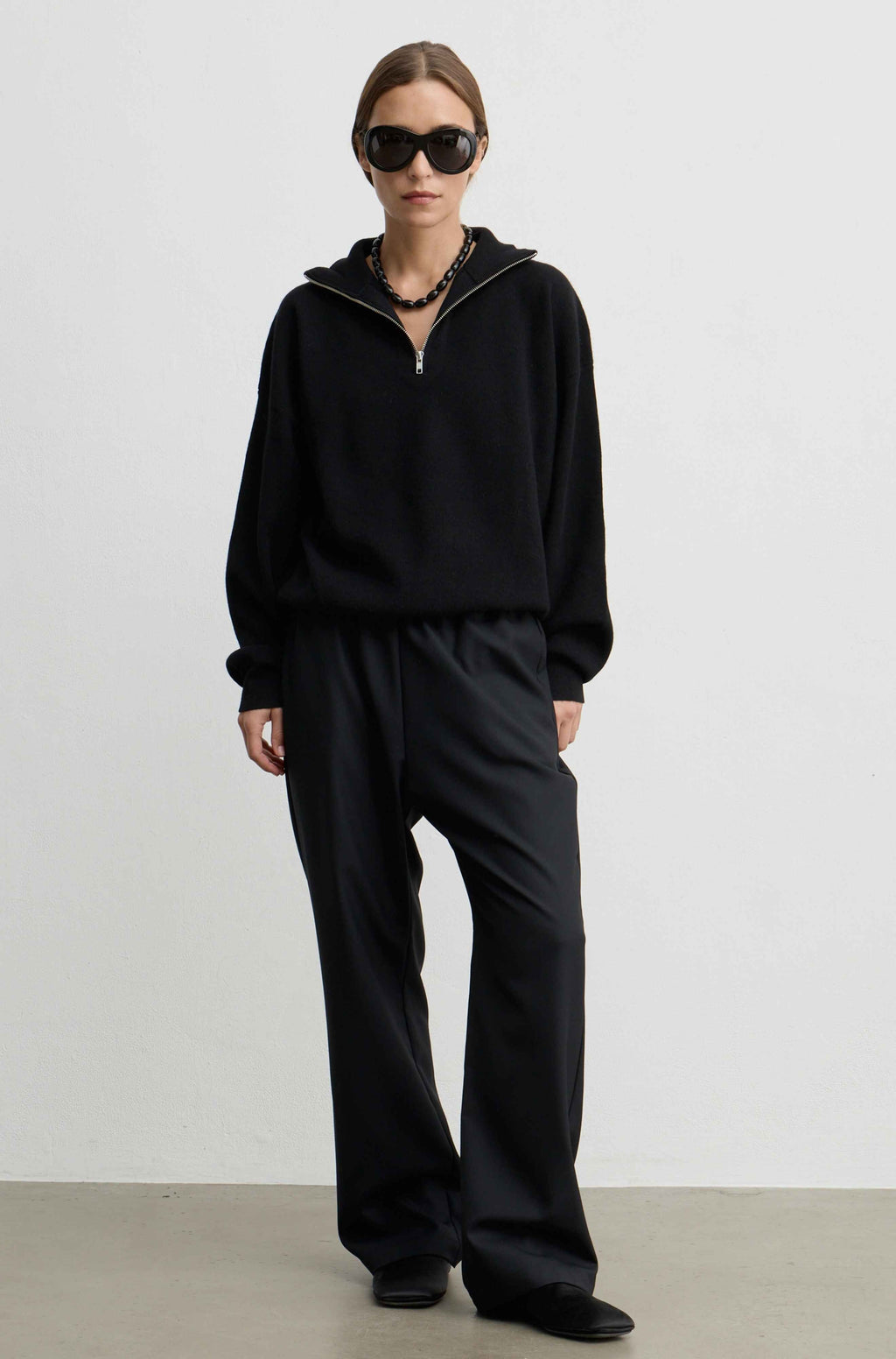 A model wearing a black half-zip hoodie and matching black wide-leg trousers, styled with black sunglasses, a chunky black necklace, and black shoes, standing against a plain light gray studio background.