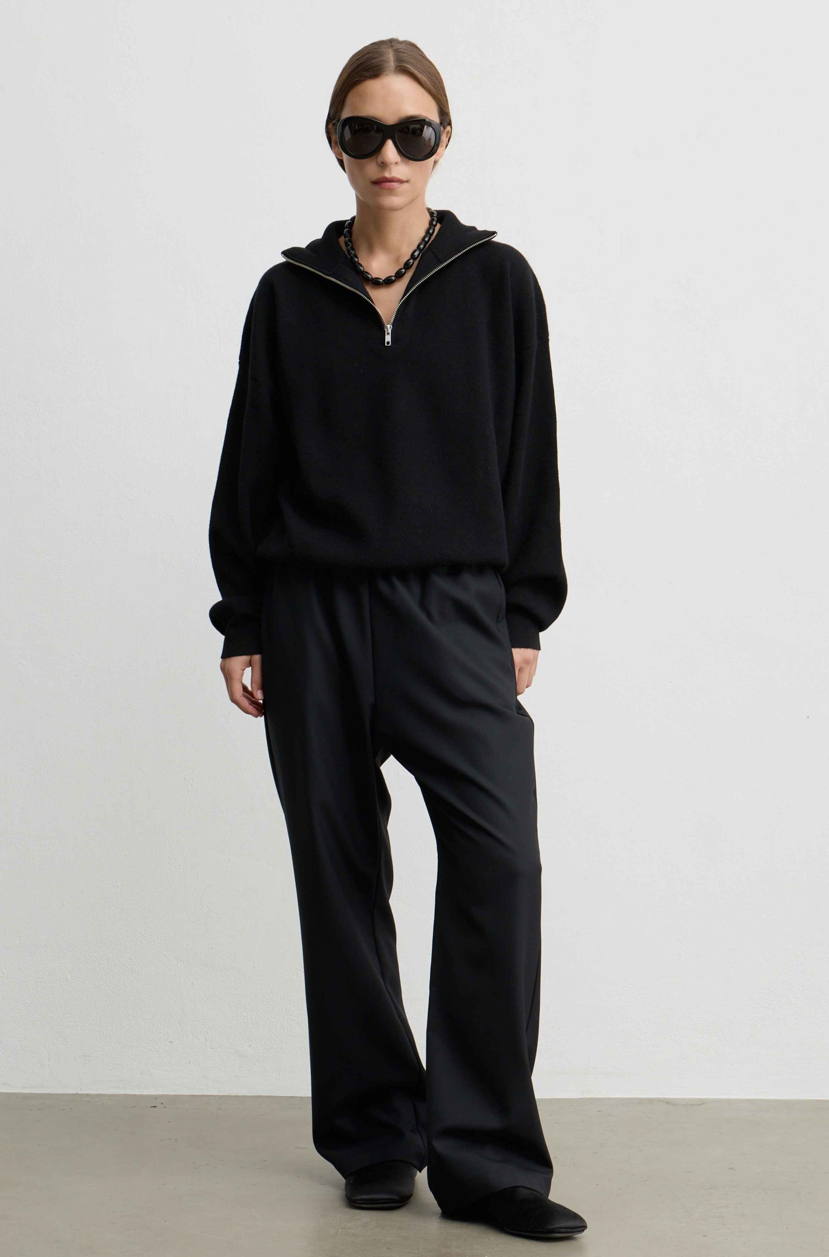 A model wearing a black half-zip hoodie and matching black wide-leg trousers, styled with black sunglasses, a chunky black necklace, and black shoes, standing against a plain light gray studio background.