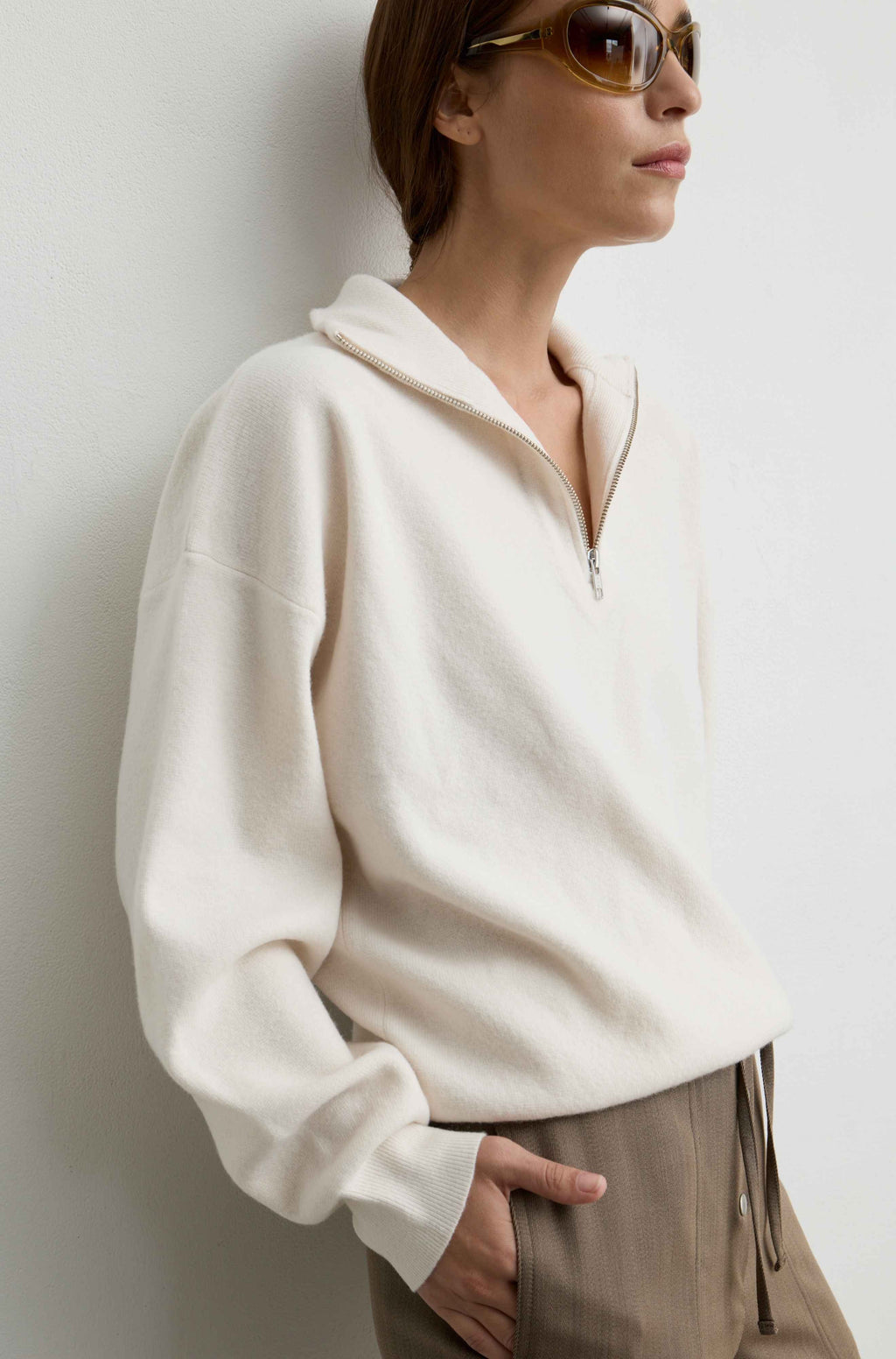 A woman wearing a cream-colored, half-zip pullover sweater and beige trousers, leaning against a light gray wall.