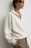 A woman wearing a cream-colored, half-zip pullover sweater and beige trousers, leaning against a light gray wall.
