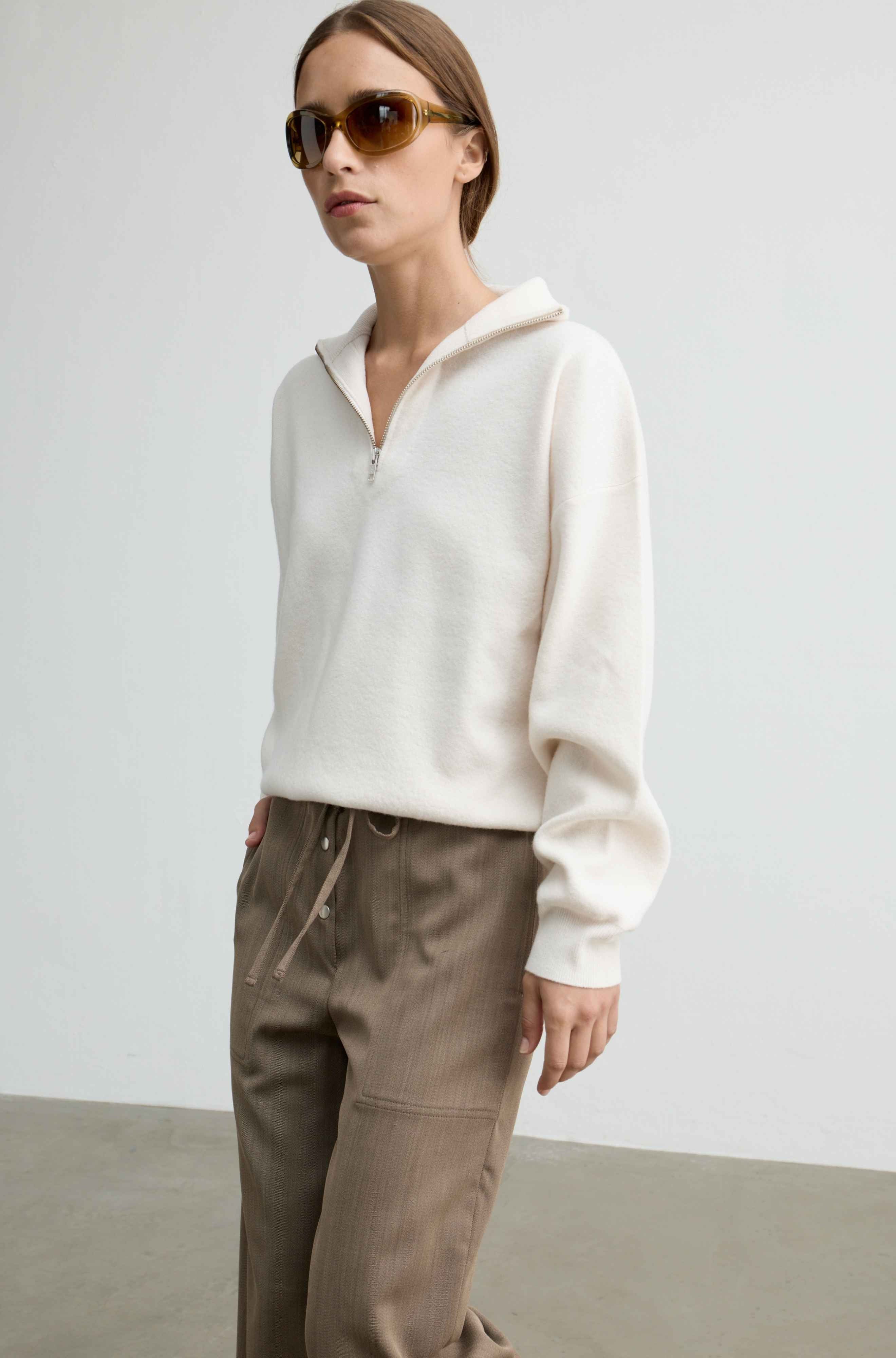 A woman wearing a cream-colored half-zip pullover sweater and brown drawstring trousers, styled with tortoiseshell sunglasses, standing against a minimalist light gray studio background.