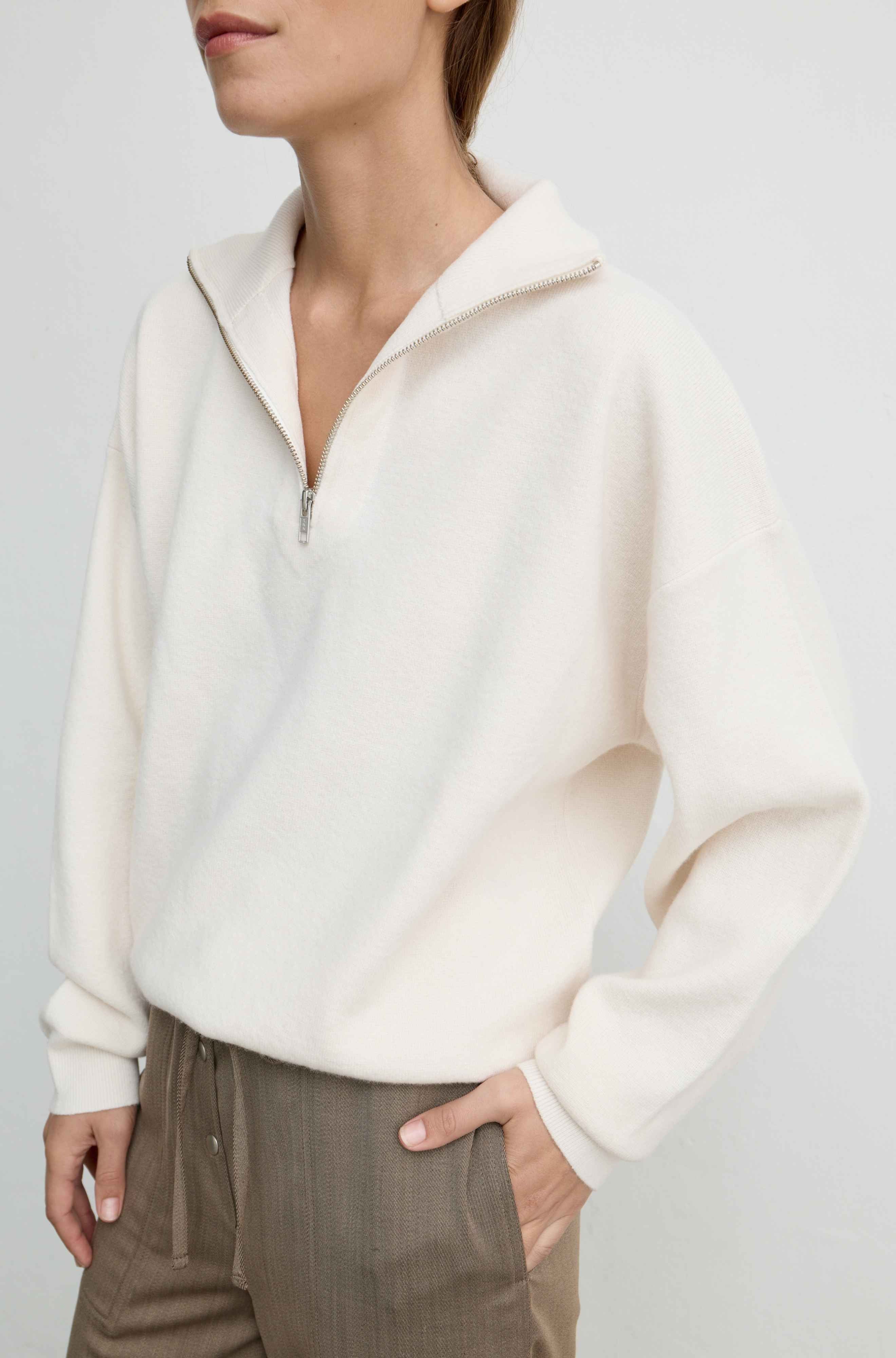 Woman wearing a zip knit sweater in cream with brown pants against a white background