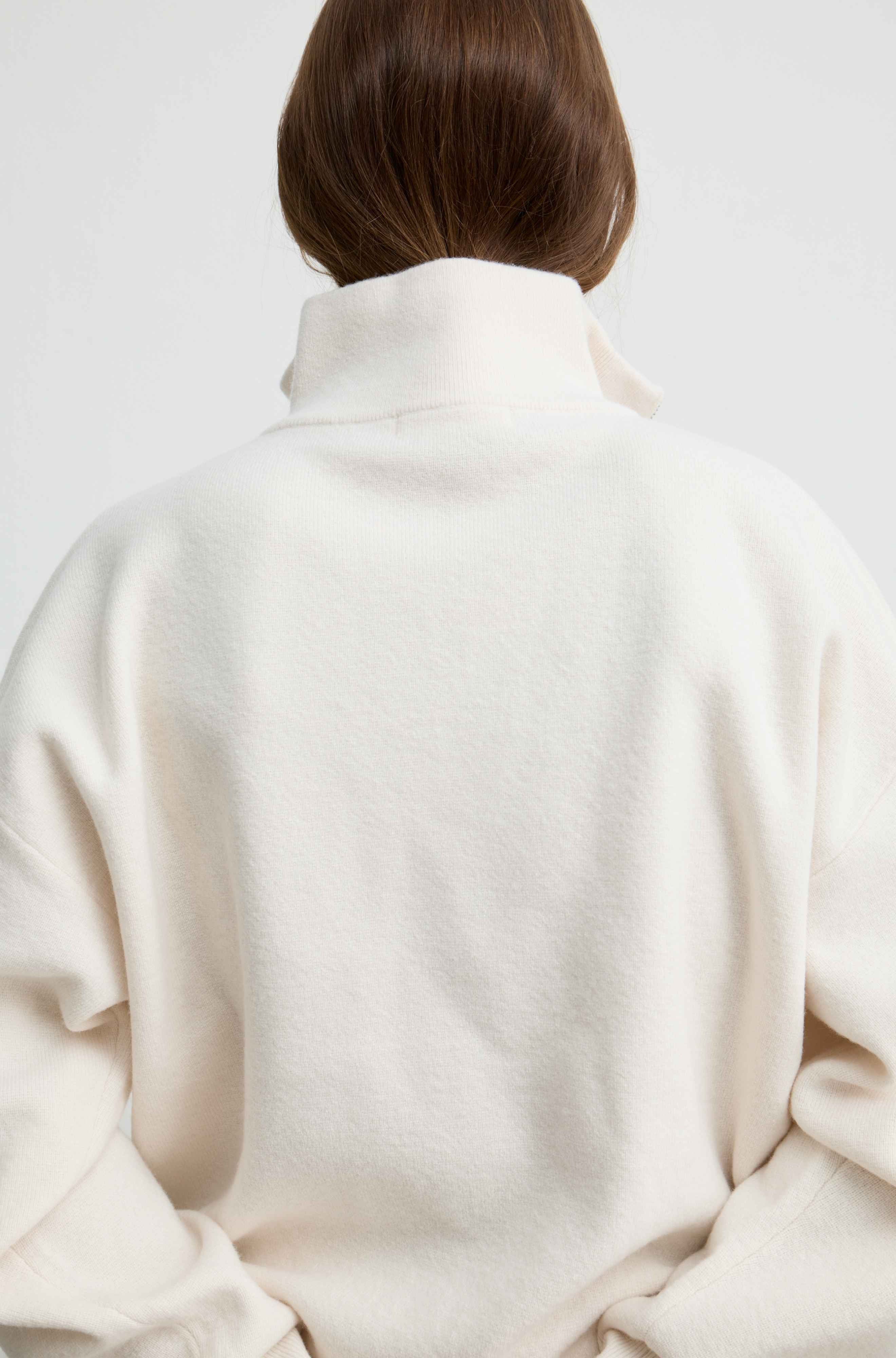 Back view of a person wearing an oversized cream-colored turtleneck sweater against a plain light gray background.