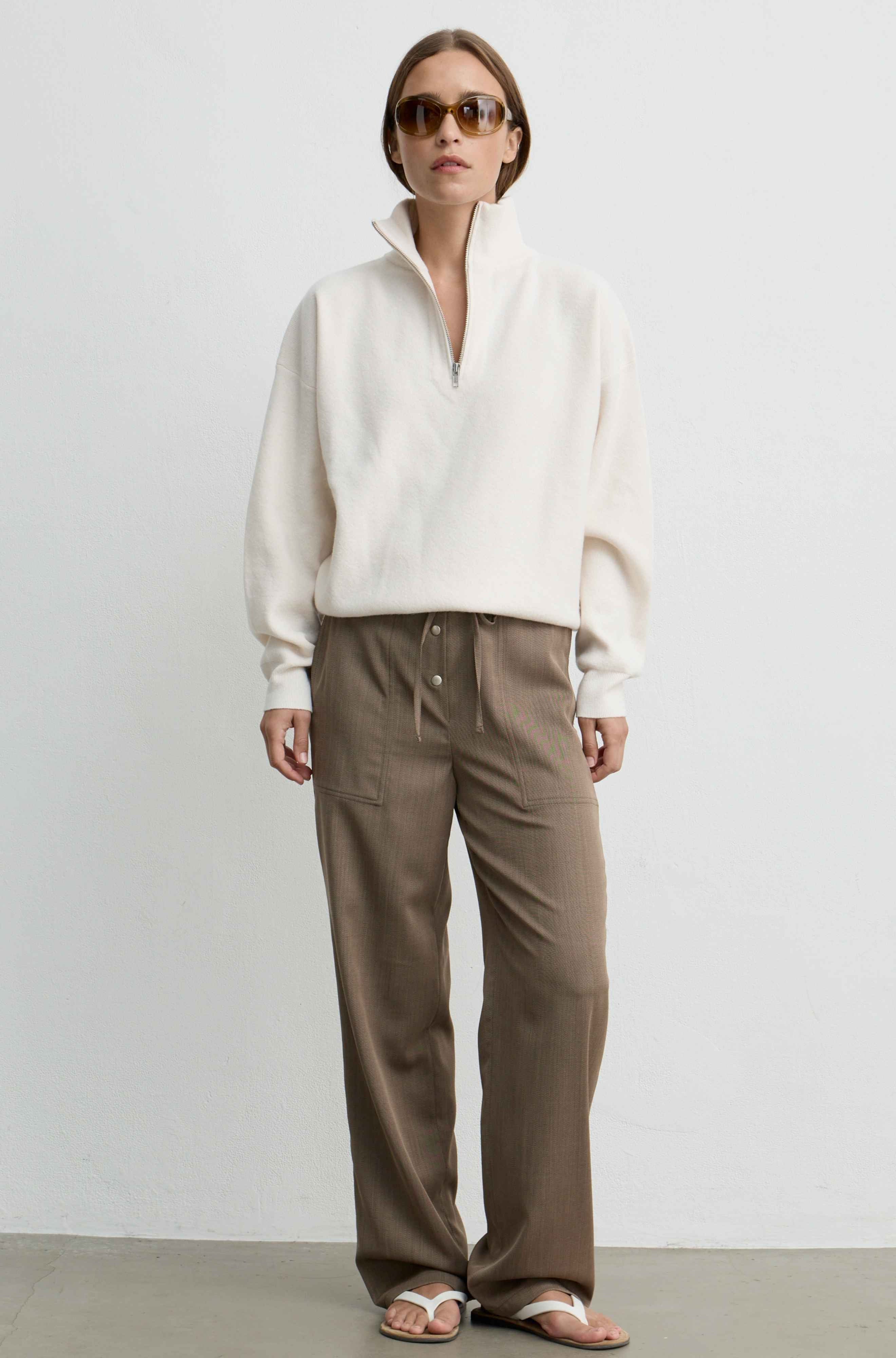 A model stands against a plain light gray wall wearing a cream-colored quarter-zip mock-neck sweater, brown wide-leg trousers, and brown sandals.