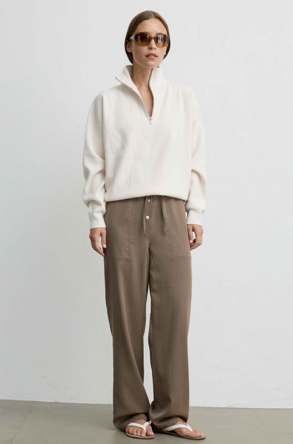A woman stands against a plain light gray wall wearing a cream half-zip pullover, brown wide-leg drawstring pants, and white sandals.
