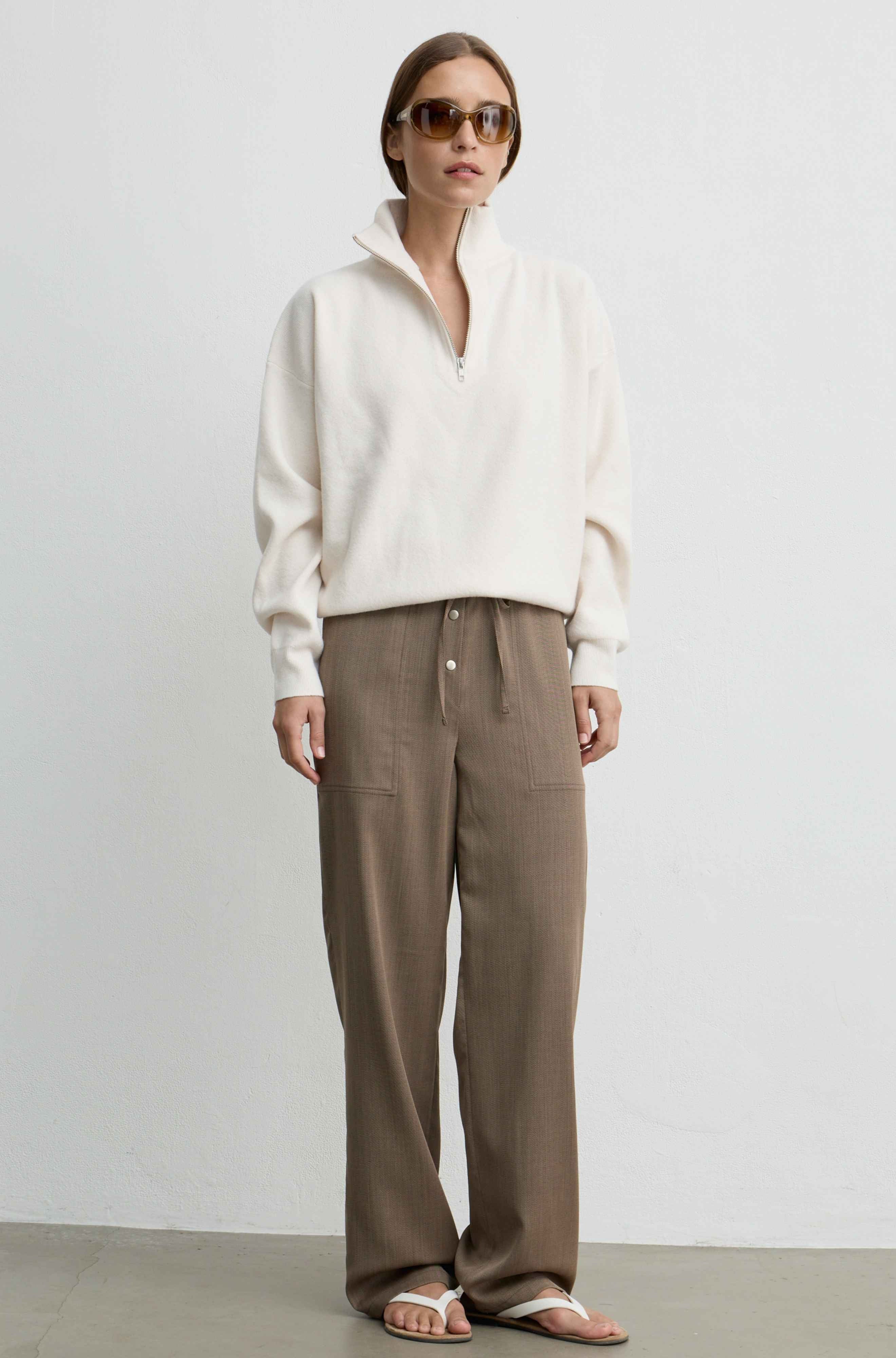 A woman stands against a plain light gray wall wearing a cream half-zip pullover, brown wide-leg drawstring pants, and white sandals.