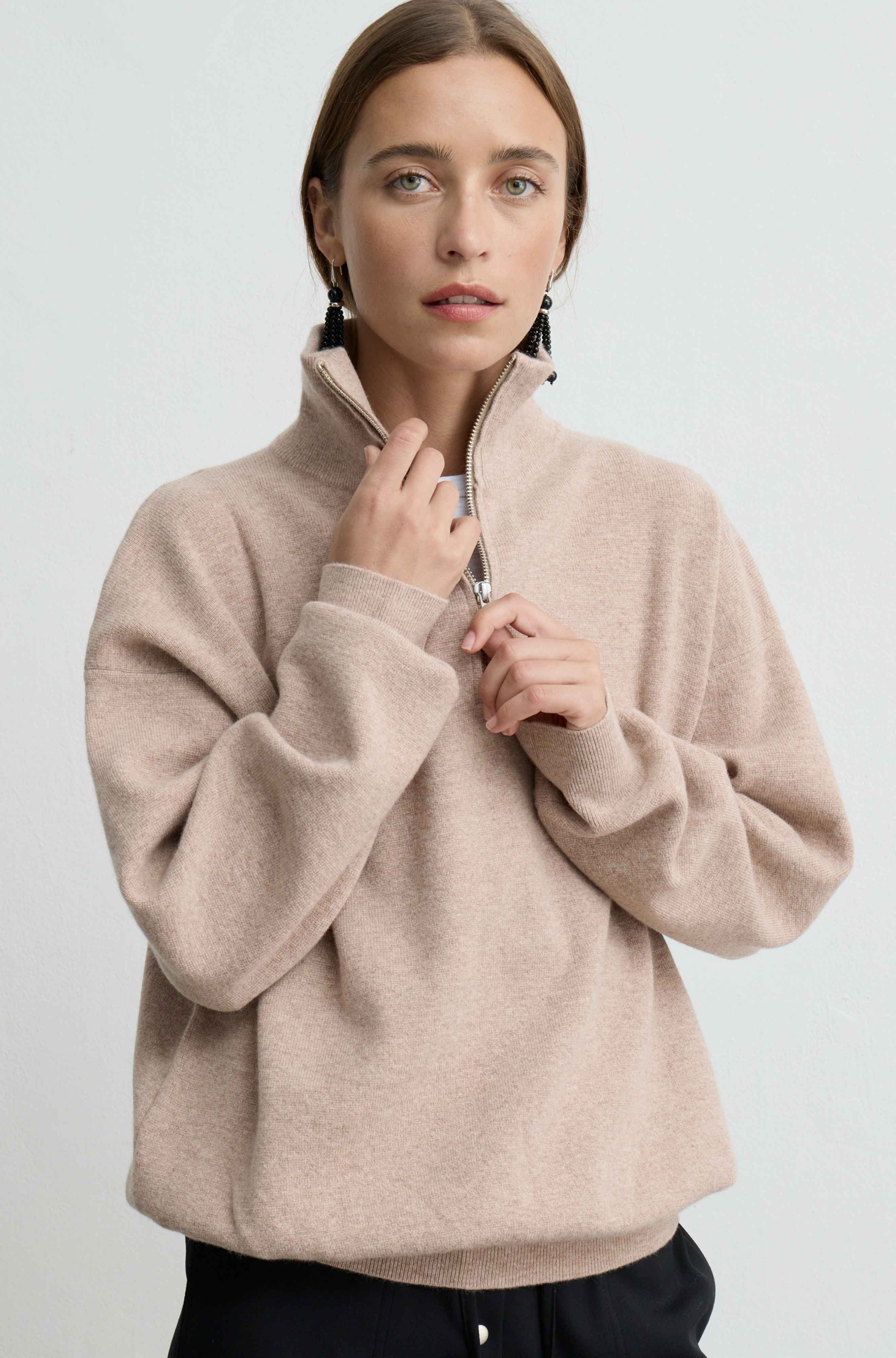 A woman wearing a light beige, high-collar quarter-zip pullover sweater, shown from the waist up against a plain light gray studio background.