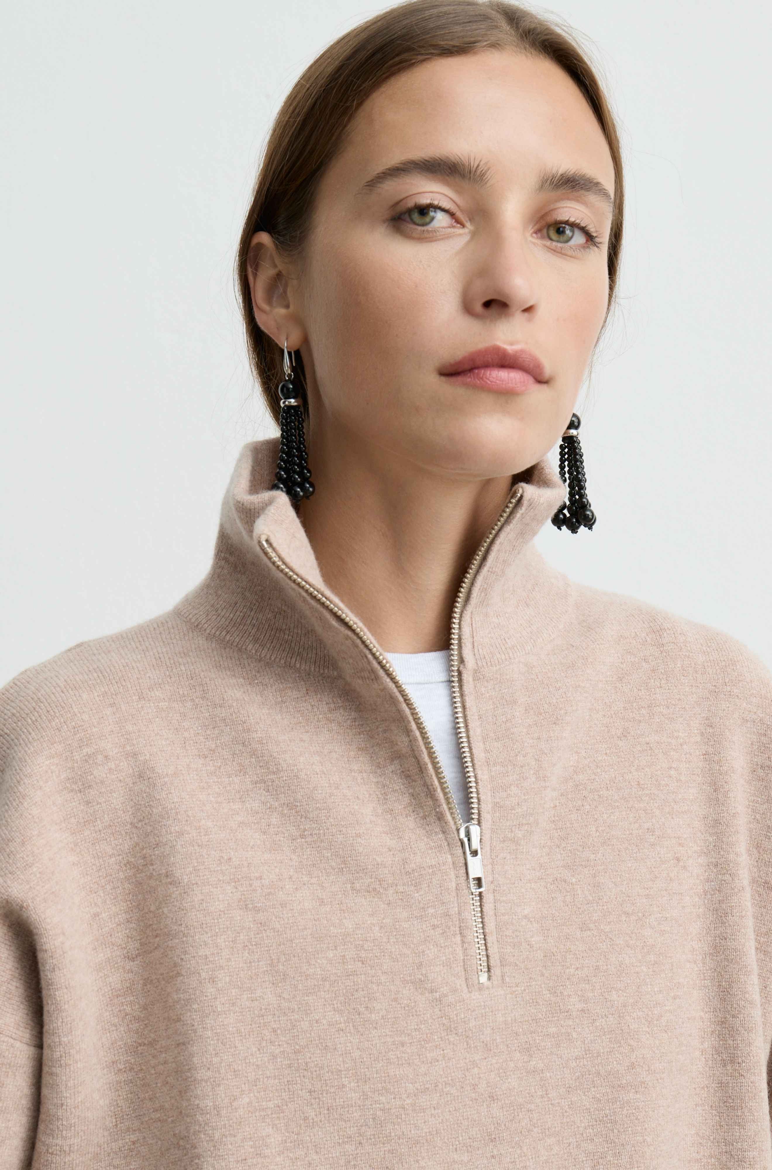 A close-up of a woman wearing a light beige quarter-zip pullover sweater over a white undershirt, styled with long black beaded drop earrings against a light gray background.