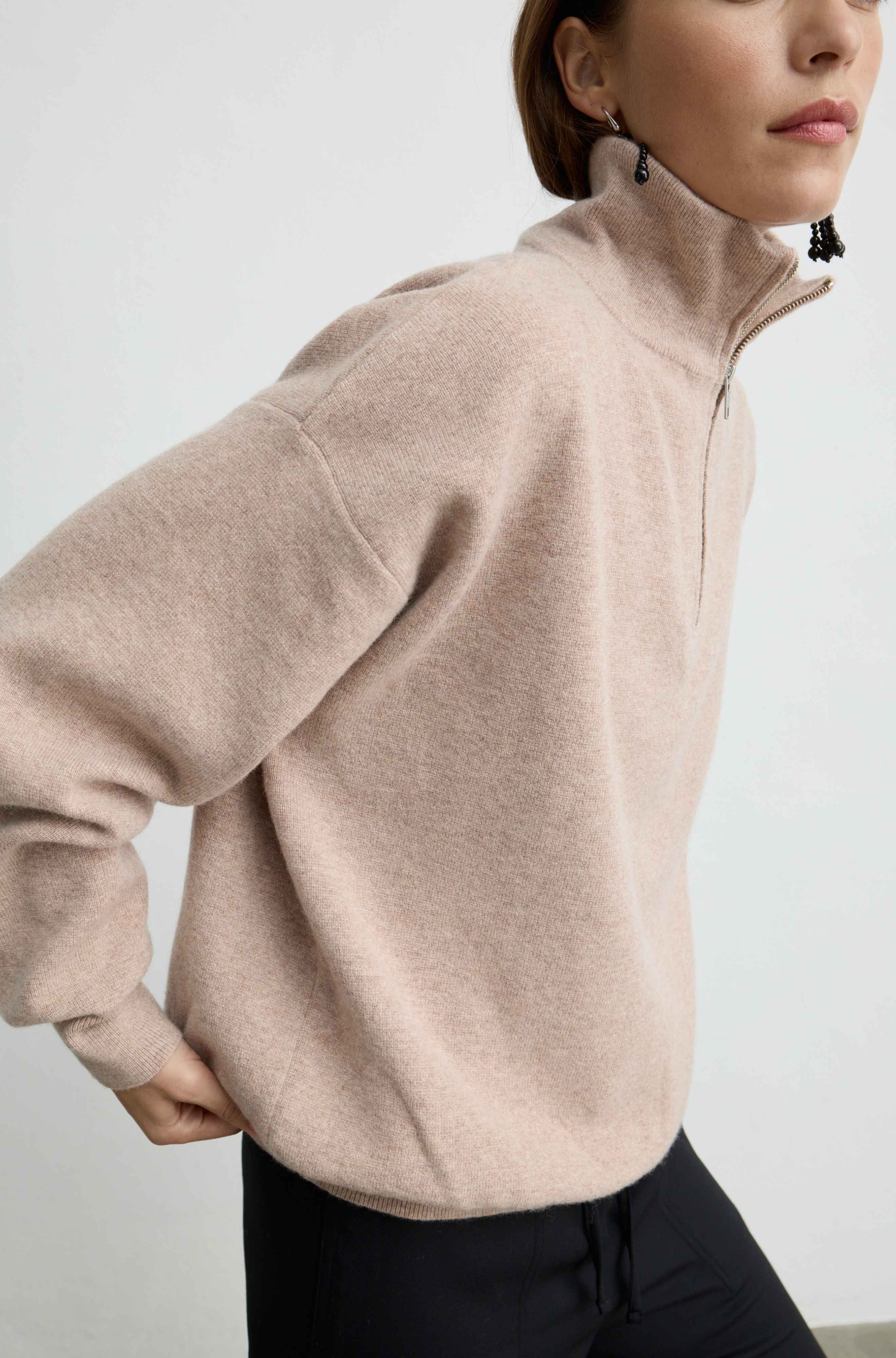 A woman wearing a light beige high-neck quarter-zip sweater and black pants, shown from a side profile against a plain light gray background.
