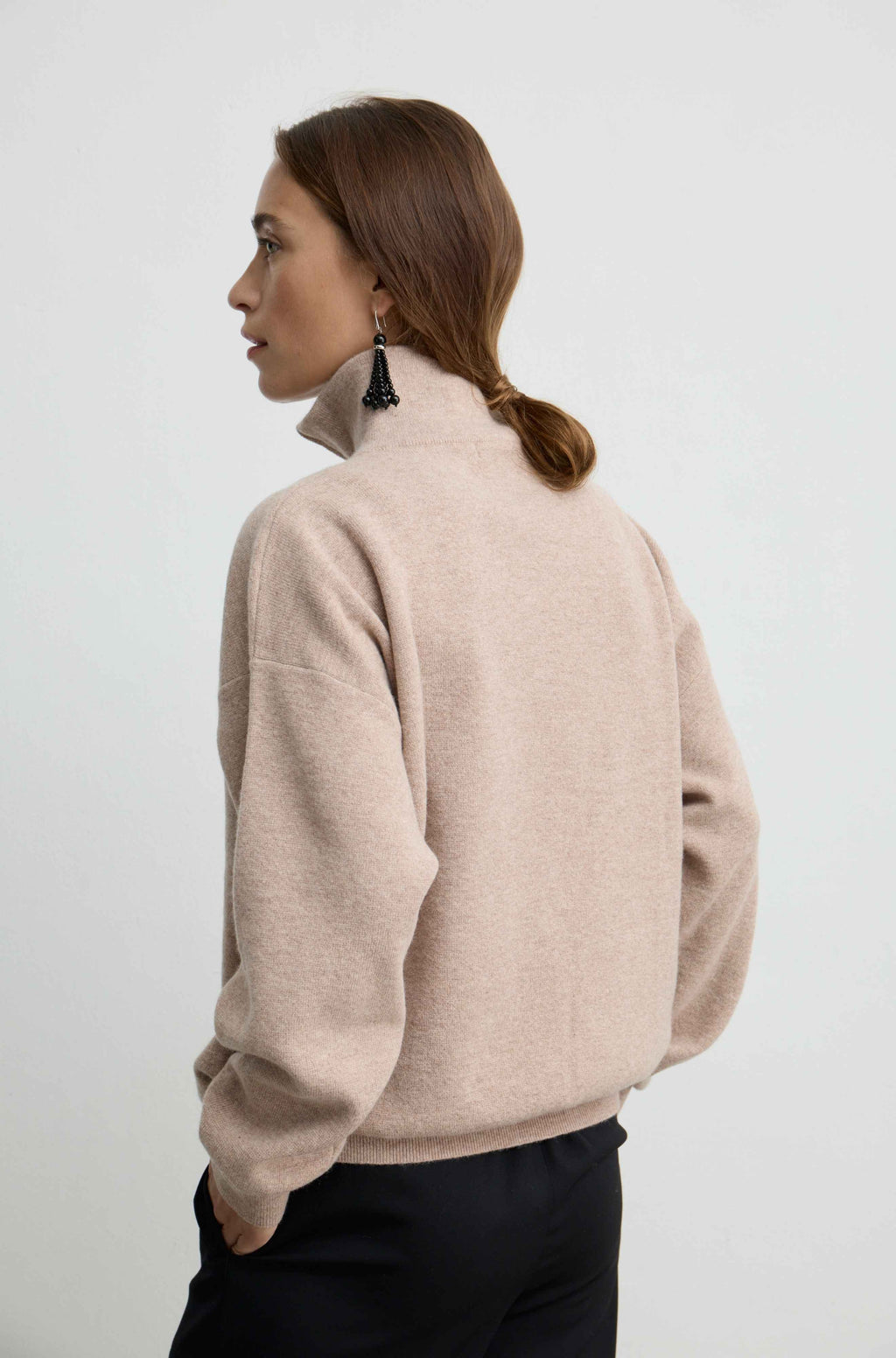 A woman shown from the back wearing a light beige knit sweater with a mock neck and black trousers, photographed against a plain light gray studio background.