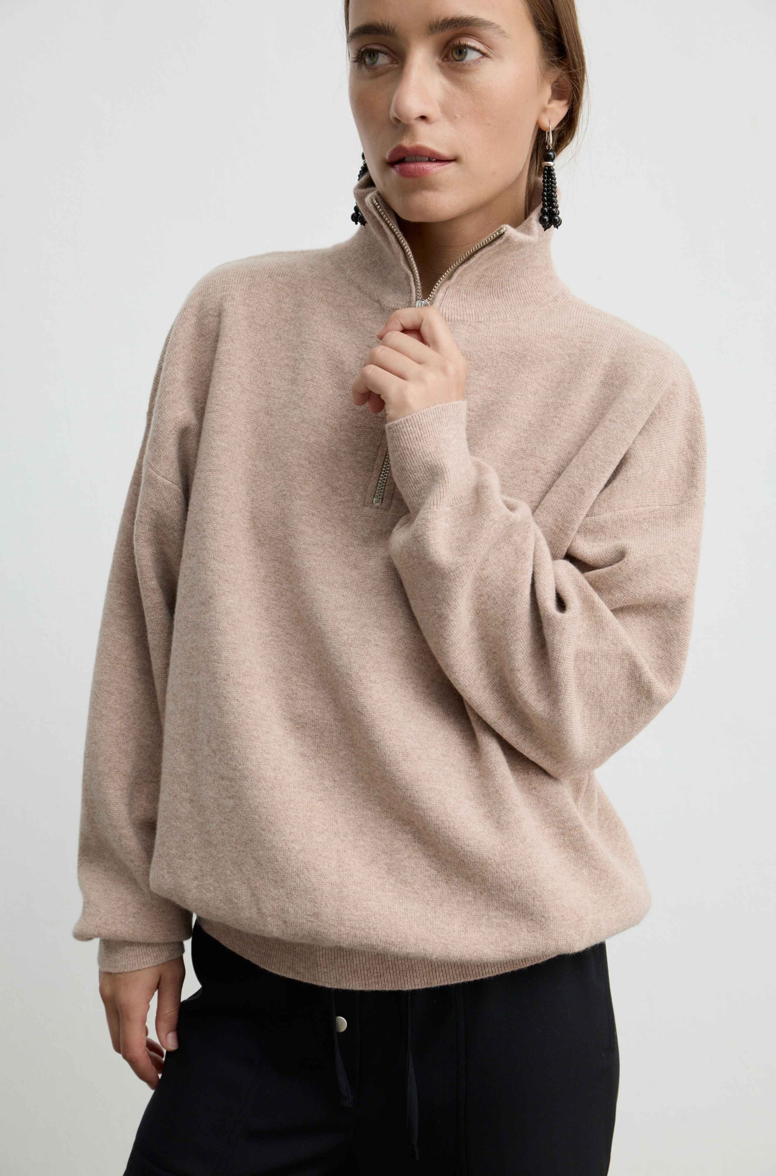 A woman wearing a light beige, quarter-zip knit sweater with a mock neck and long sleeves, paired with black pants, against a plain light gray background.