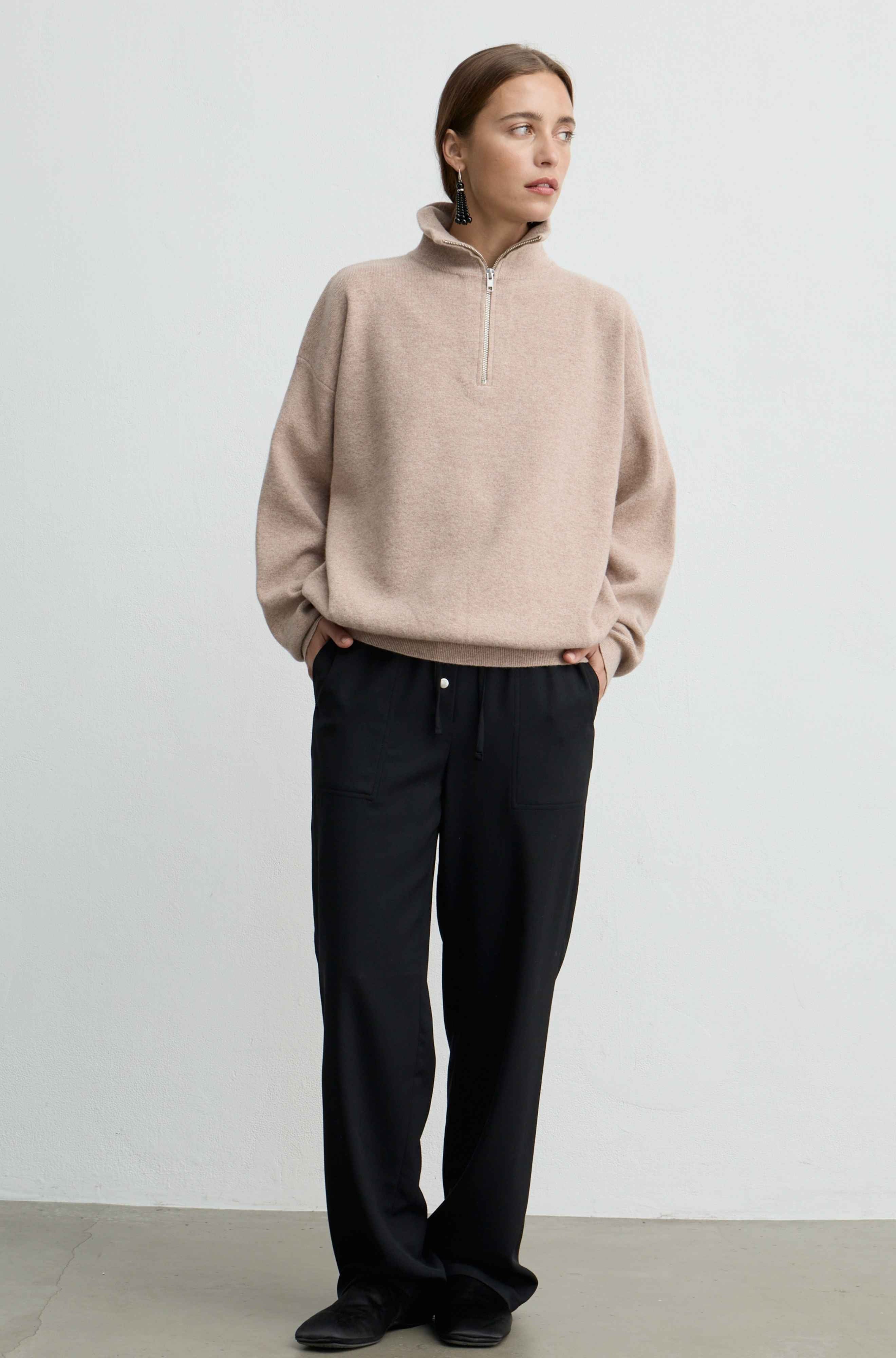 A woman modeling a light beige quarter-zip pullover sweater and black wide-leg trousers against a plain light gray studio background.