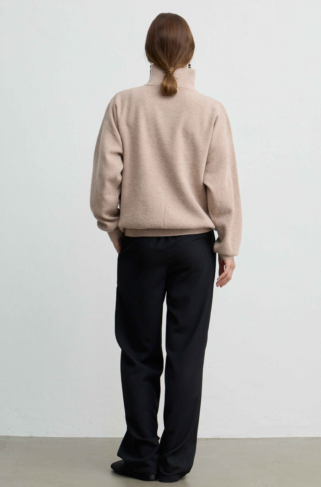 A model viewed from behind wearing a light beige mock-neck knit sweater and black wide-leg trousers, standing against a plain white wall.