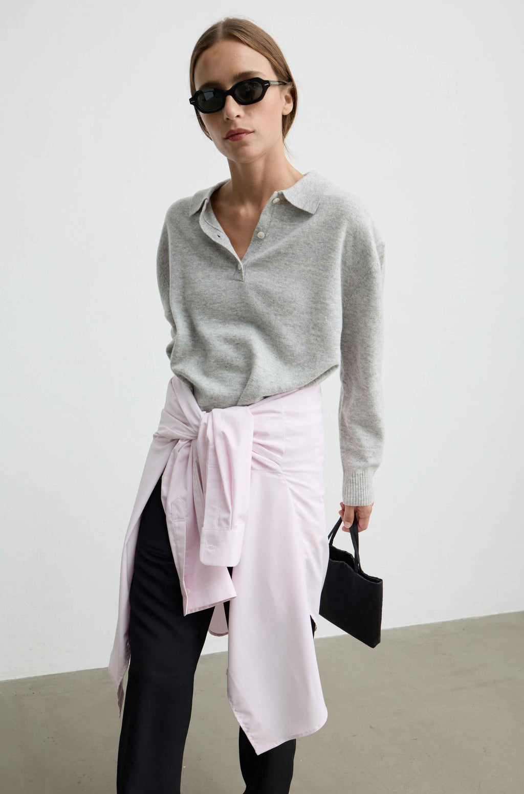 A woman modeling a light gray knit polo shirt, styled with a light pink wrap skirt tied at the waist, black pants, black sunglasses, and holding a small black handbag against a minimalist studio background.