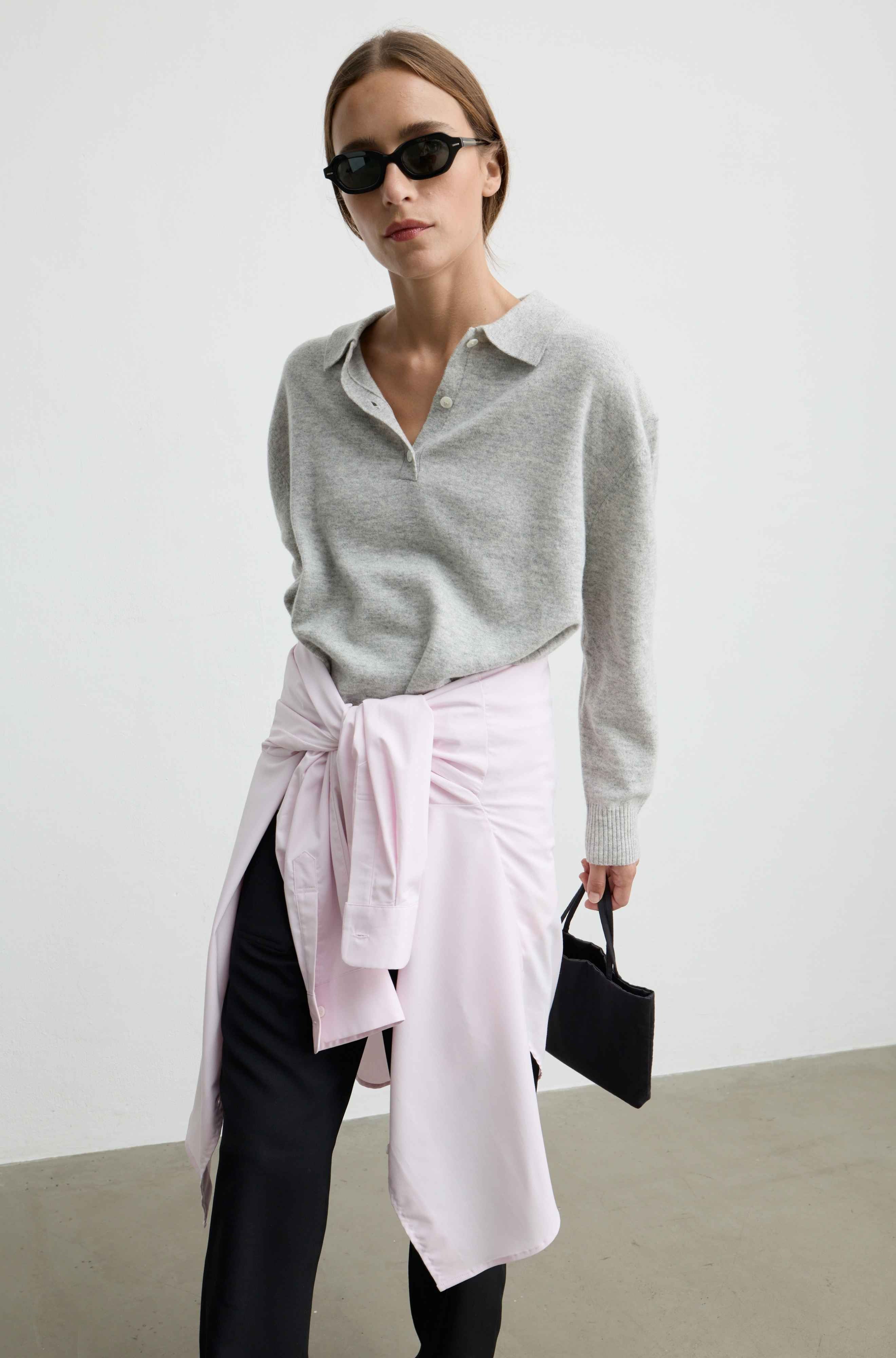 A woman modeling a light gray knit polo shirt, styled with a light pink wrap skirt tied at the waist, black pants, black sunglasses, and holding a small black handbag against a minimalist studio background.