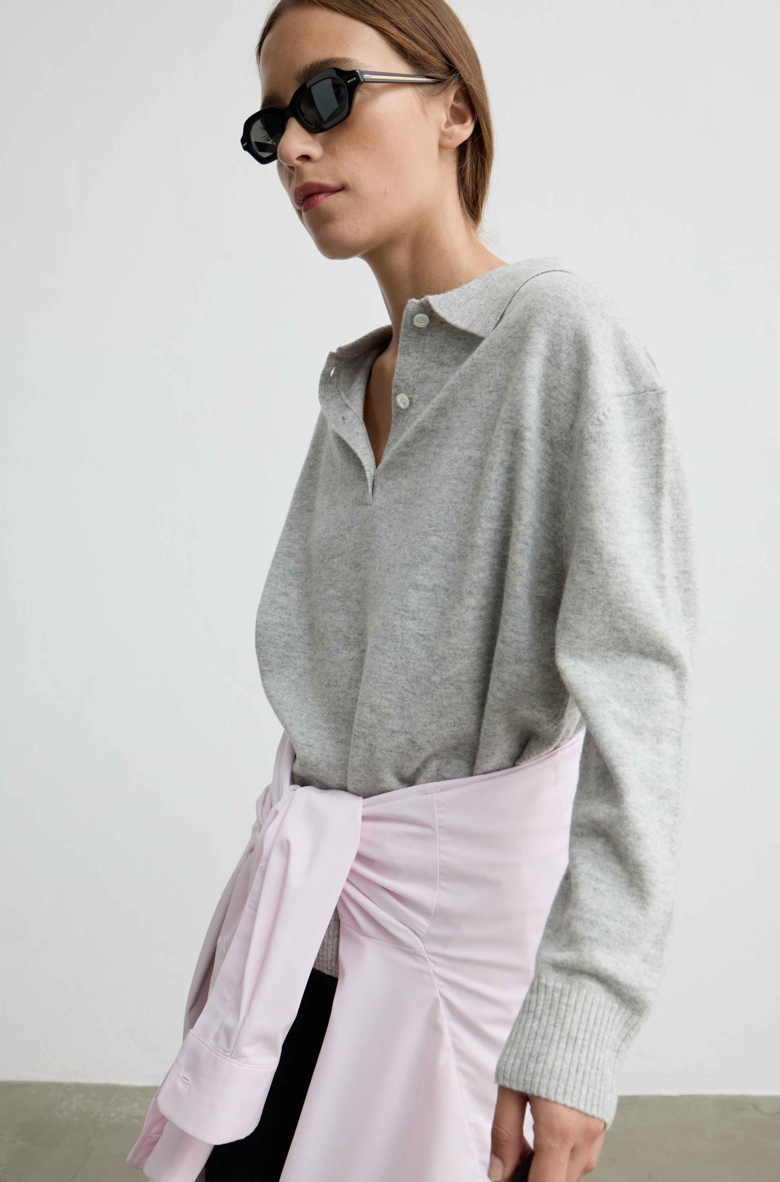 A woman wearing a light gray knit polo-style sweater and a light pink draped wrap skirt, photographed against a plain light gray wall.