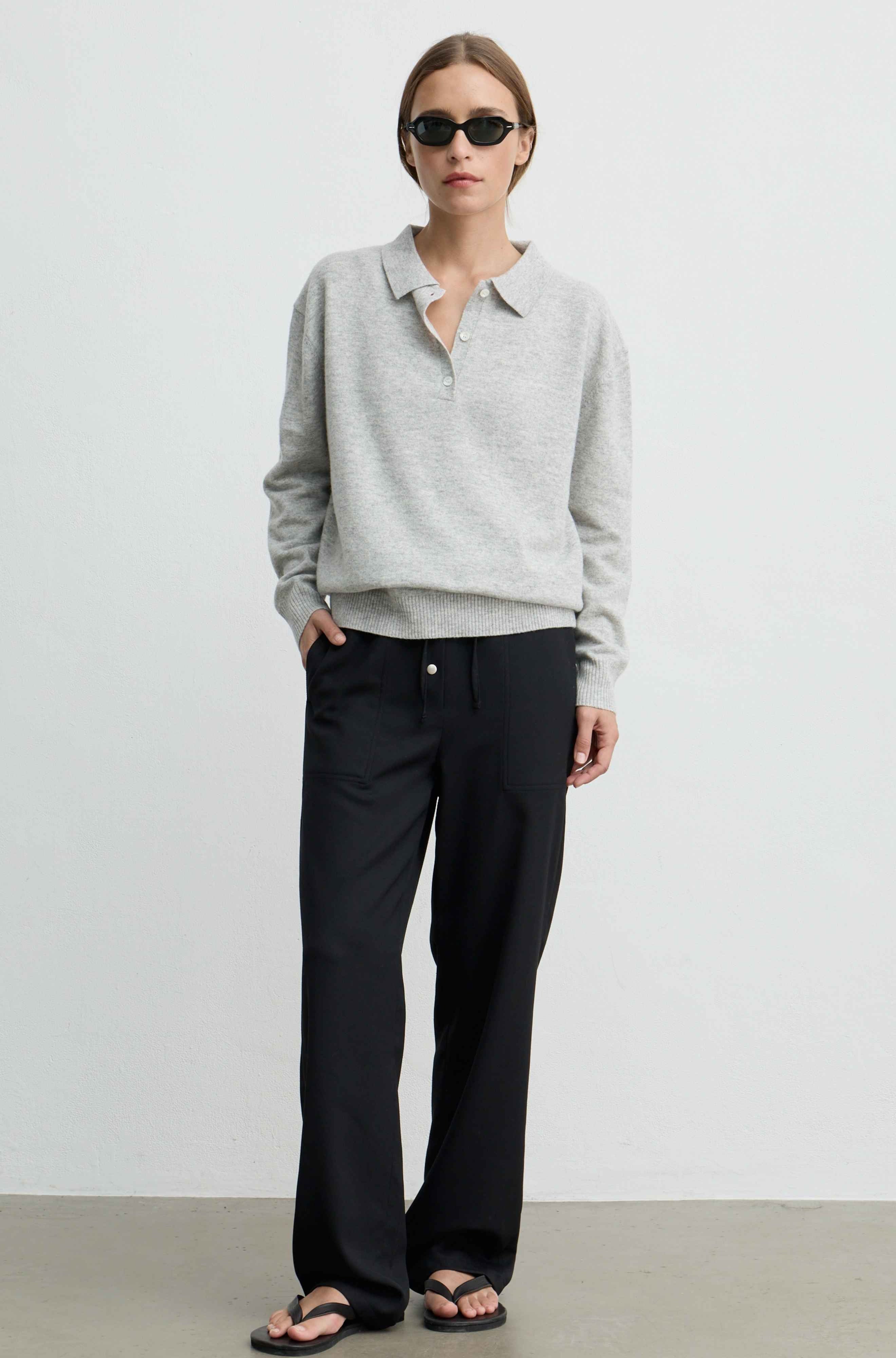 Woman wearing a light grey wool poloknit sweater with black pants, black sunglasses, and black sandals against a light grey background