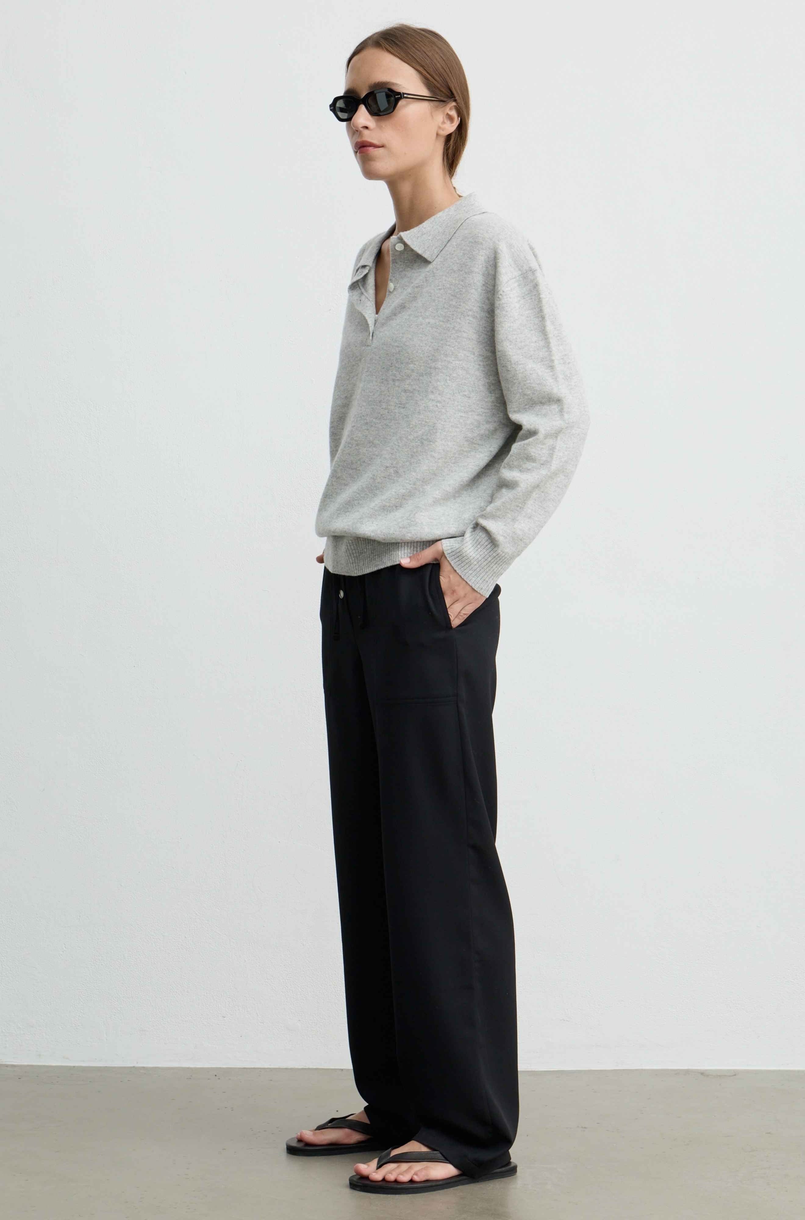 A woman stands in profile wearing a light gray long-sleeve polo sweater, black wide-leg trousers, and black sandals against a plain light gray studio background.