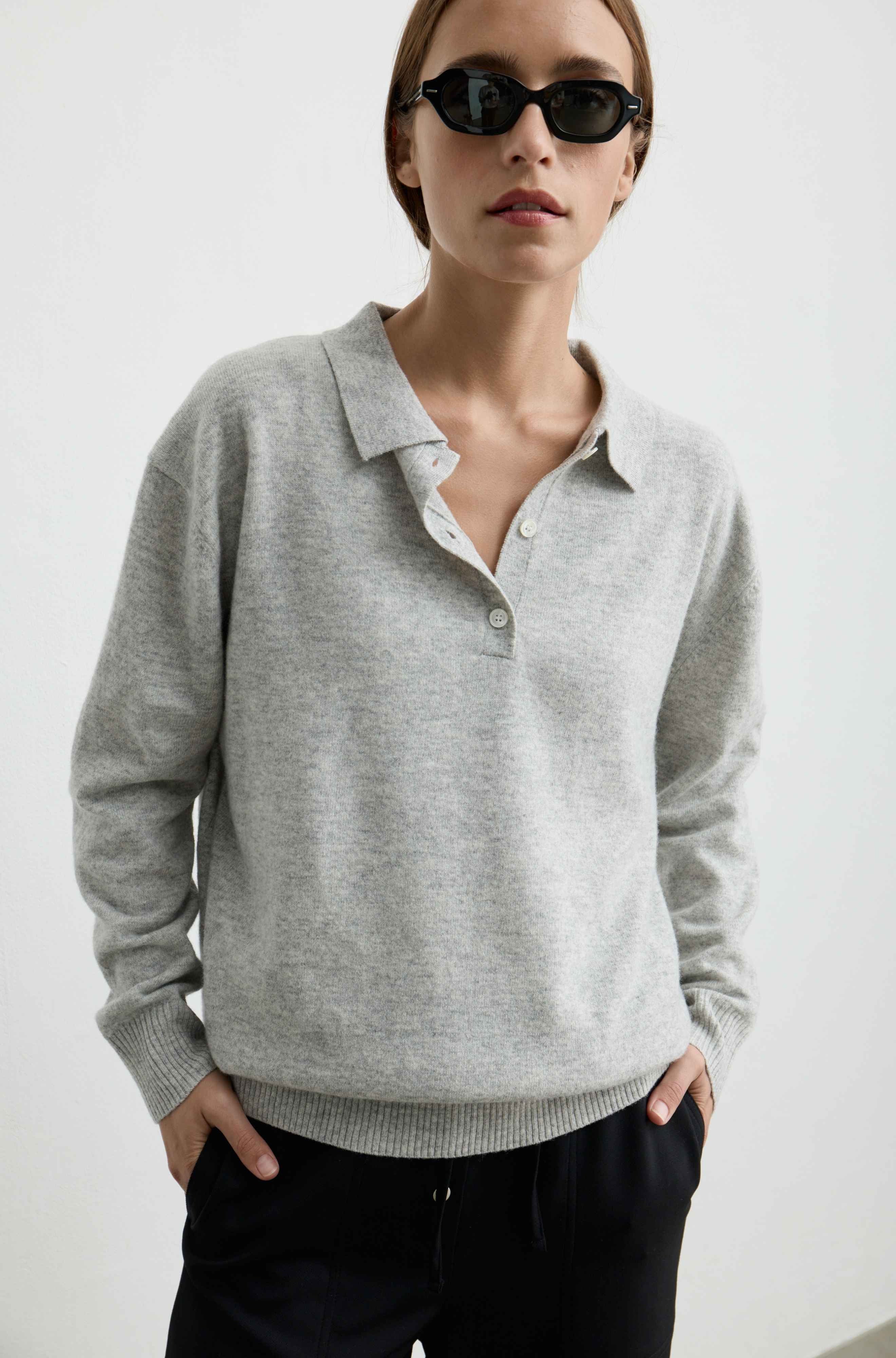 A woman wearing a light gray knit polo sweater and black pants, photographed against a plain light background.