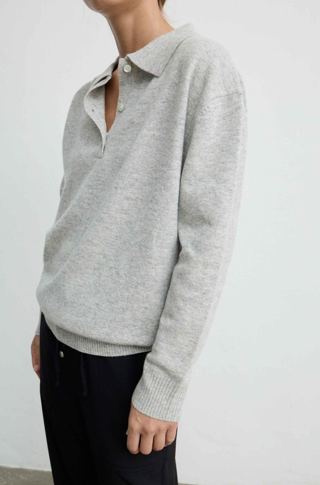 A person wearing a light heather gray knit polo sweater with a partial button placket and dark pants, photographed against a plain light gray wall.