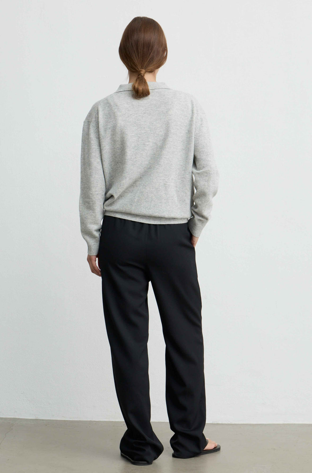 A woman viewed from behind wearing a light gray long-sleeve knit top and black wide-leg trousers, standing against a plain light gray wall.