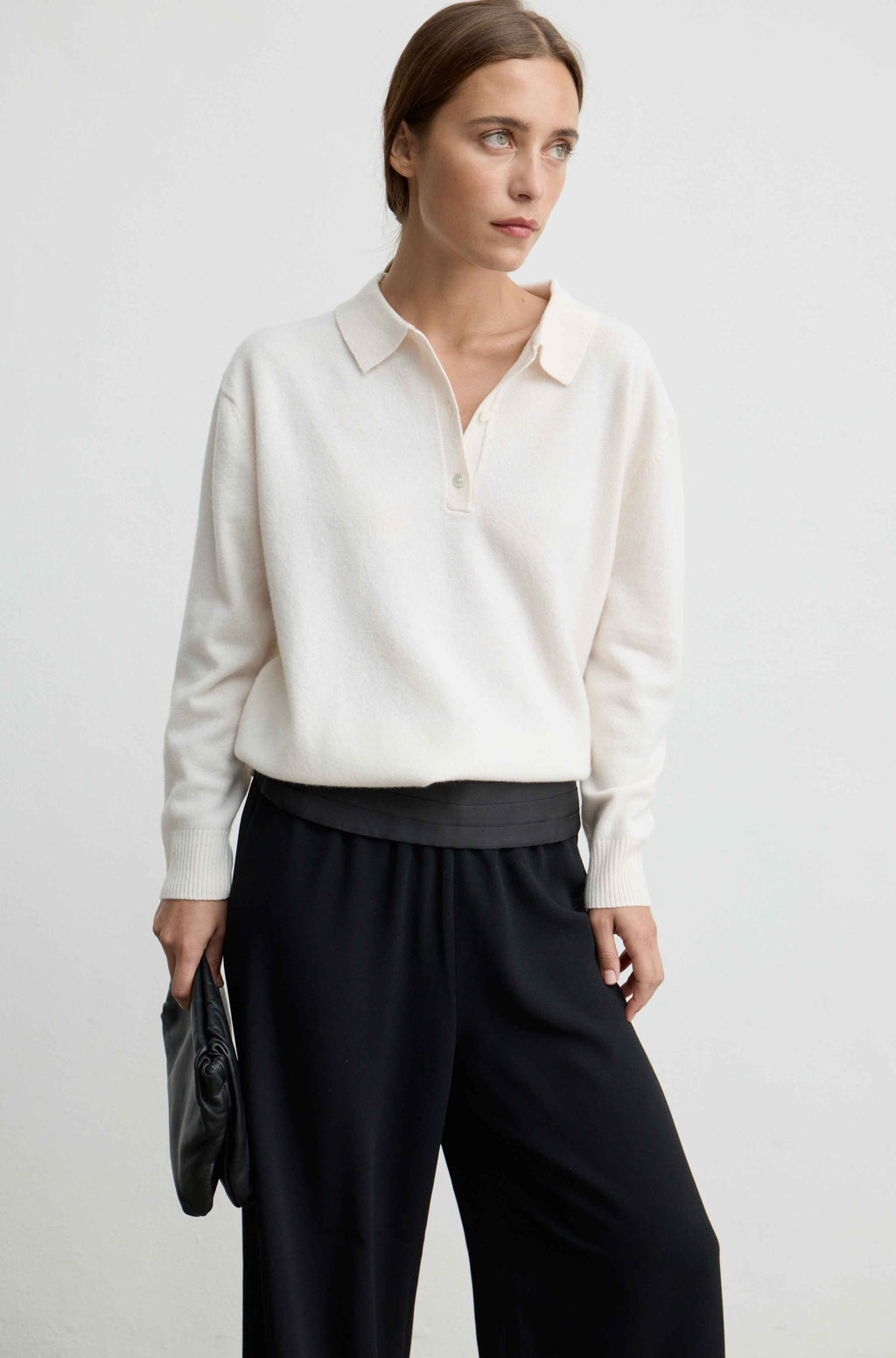 Woman wearing a white lightweight wool polo sweater with black pants and a black handbag against a light grey background