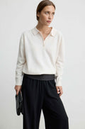 Woman wearing a white lightweight wool polo sweater with black pants and a black handbag against a light grey background