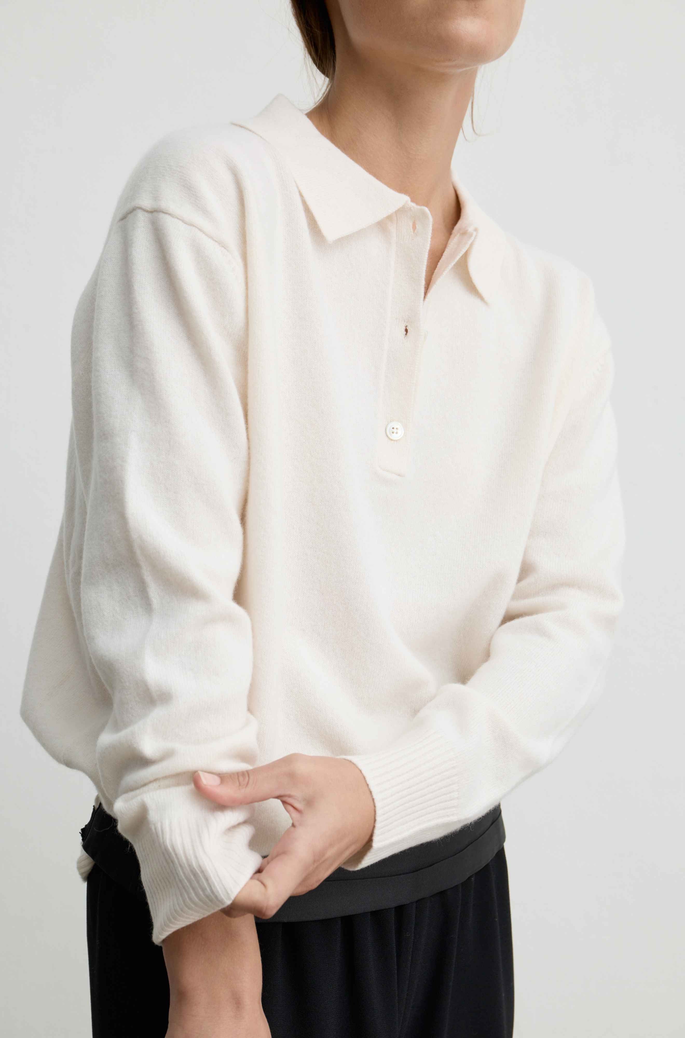 Woman wearing a white lightweight wool polo sweater with black pants against a light grey background