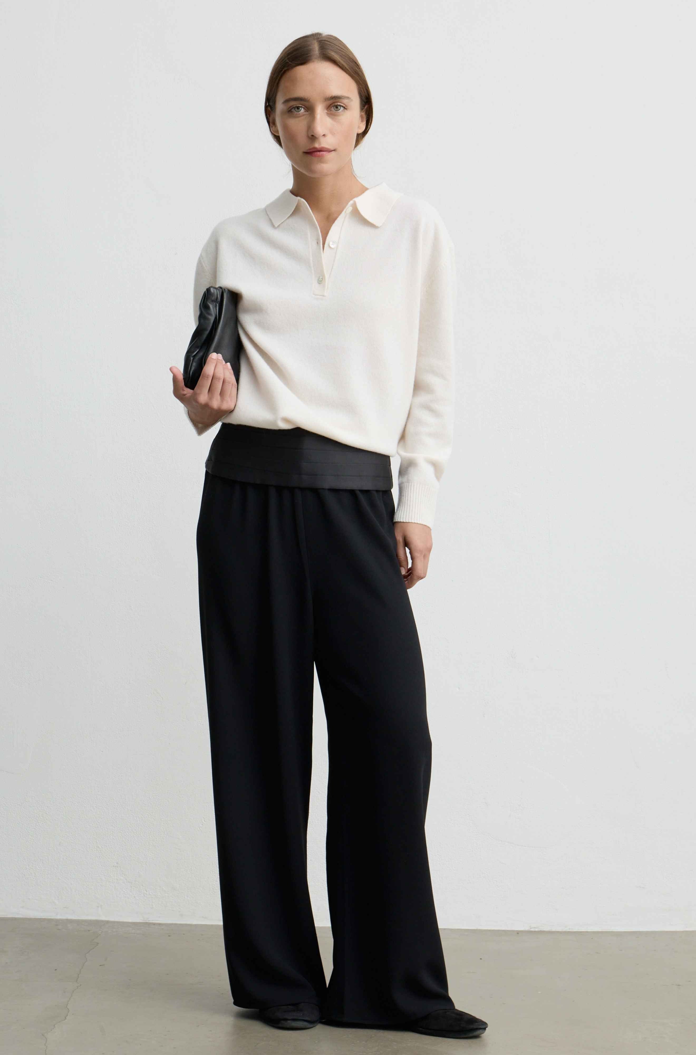 A woman stands in a studio wearing a cream long-sleeve collared top, black wide-leg trousers, and holding a small black clutch.