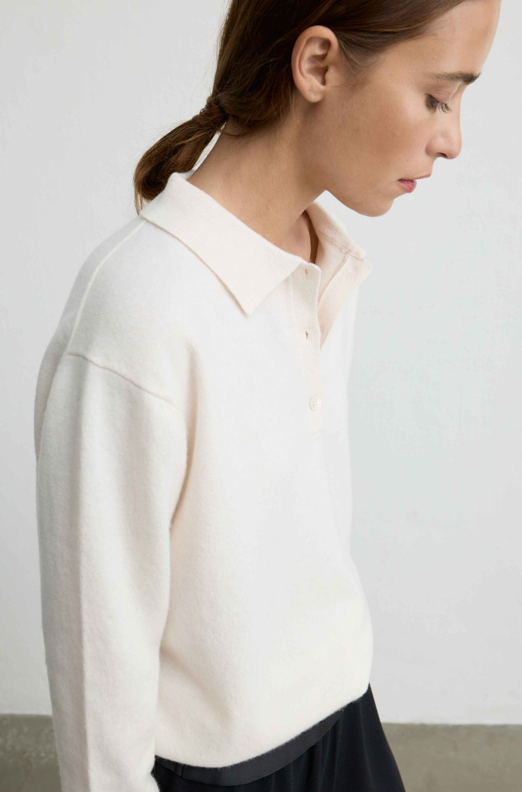 A woman shown in profile wearing a cream-colored knit polo sweater and dark pants, photographed against a plain light gray wall.