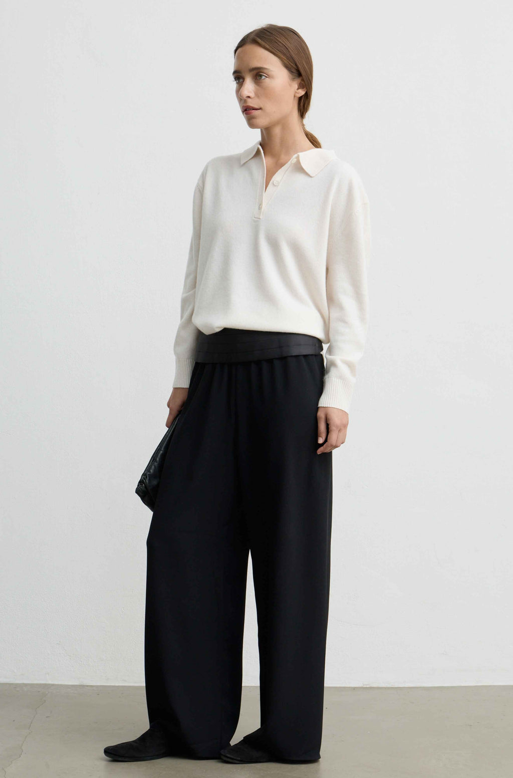A woman stands in a studio wearing a cream long-sleeve collared knit top and black wide-leg trousers, with black shoes and a black clutch.