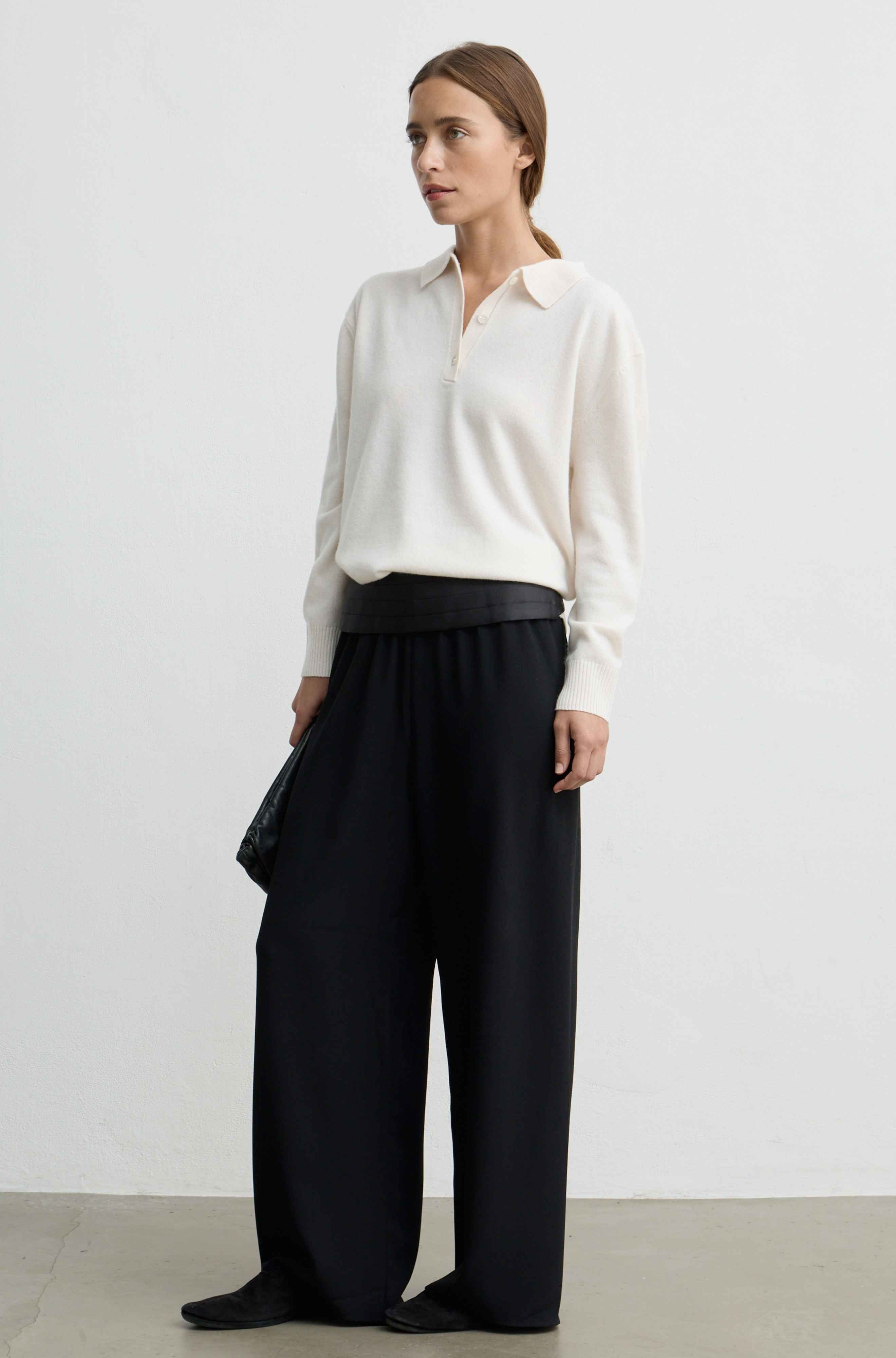 A woman stands in a studio wearing a cream long-sleeve collared knit top and black wide-leg trousers, with black shoes and a black clutch.