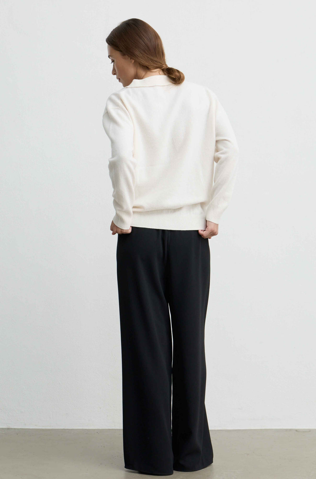 A woman shown from the back wearing a cream-colored knit sweater and black wide-leg trousers, standing against a plain light gray wall.