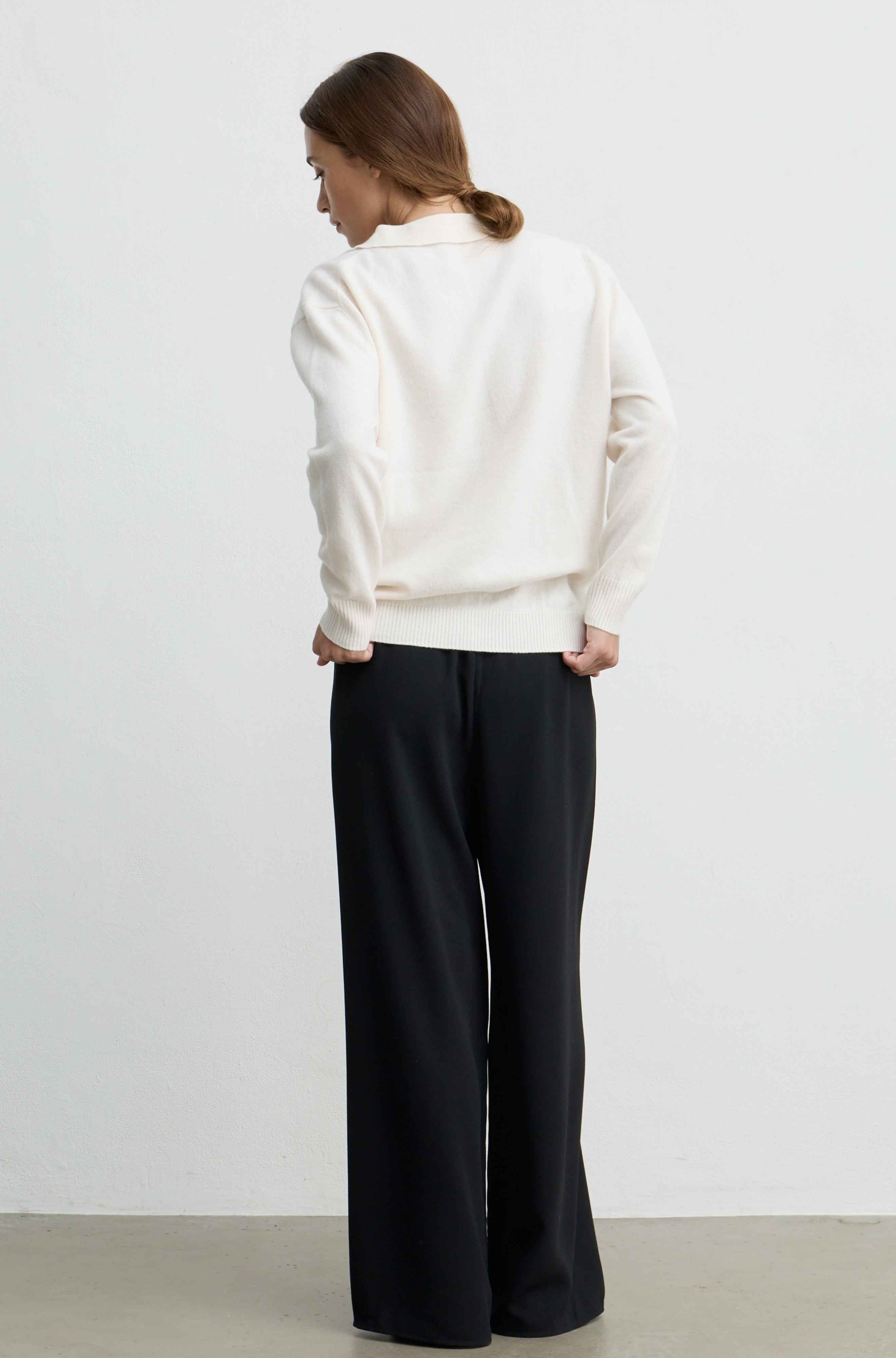 A woman shown from the back wearing a cream-colored knit sweater and black wide-leg trousers, standing against a plain light gray wall.