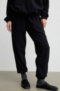 A person wearing loose black jogger-style pants with a drawstring waist, paired with gray socks and black slip-on shoes, photographed against a plain light wall and concrete floor.