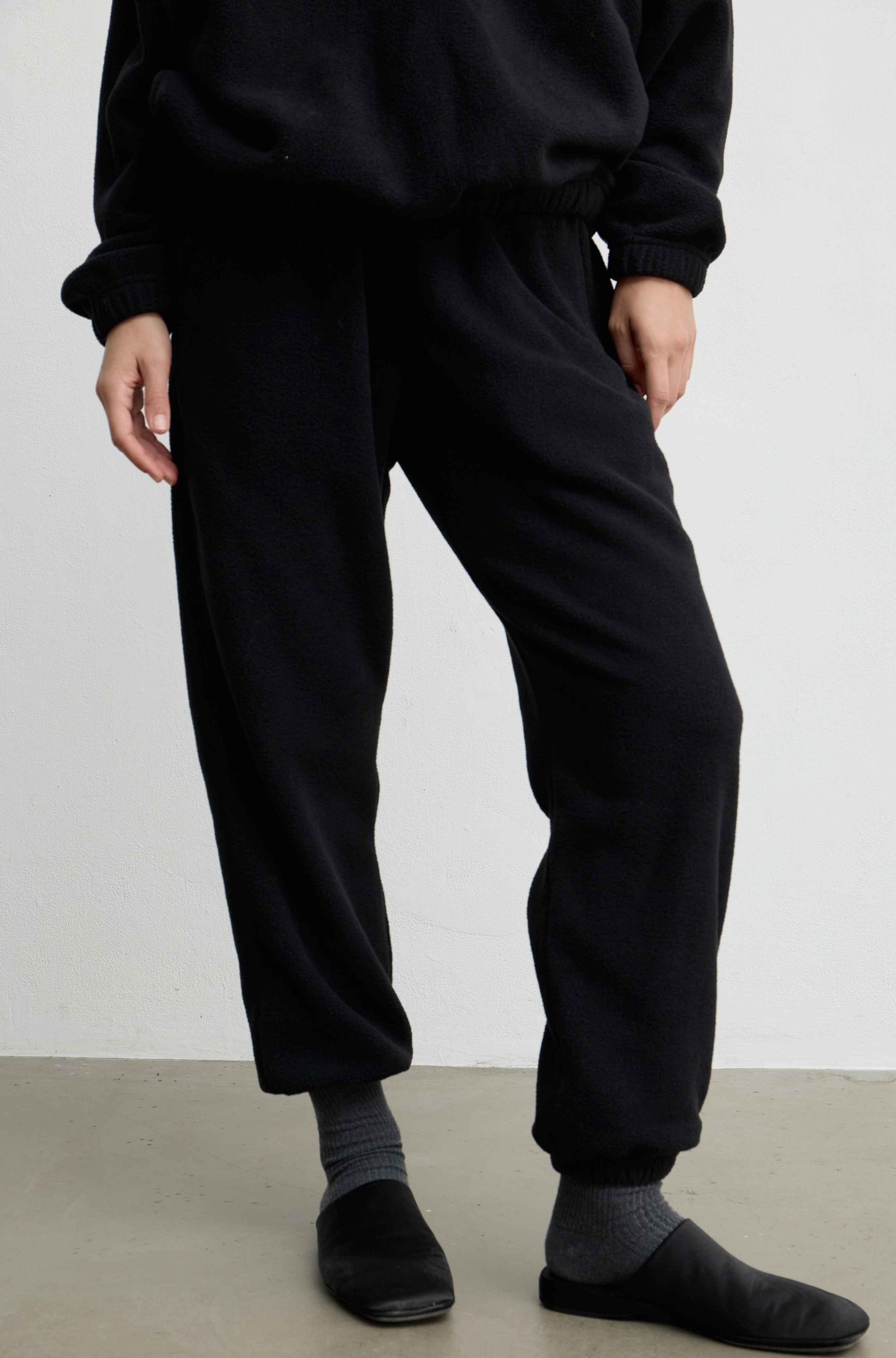 A person wearing loose black jogger-style pants with a drawstring waist, paired with gray socks and black slip-on shoes, photographed against a plain light wall and concrete floor.
