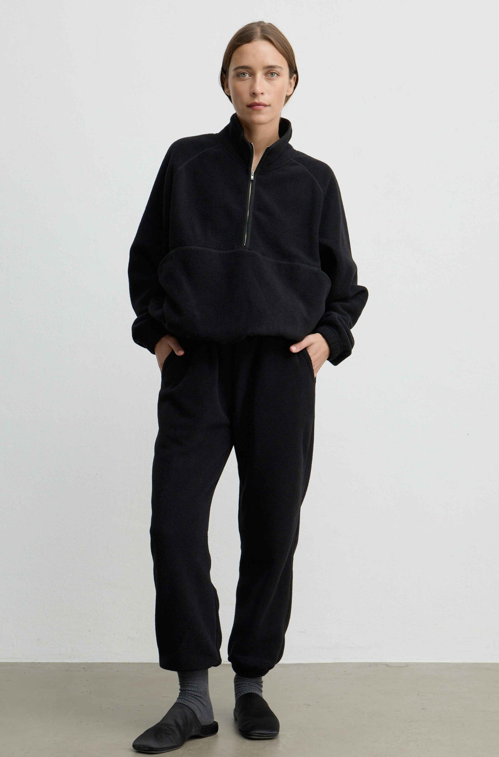 A woman modeling a black fleece half-zip pullover and matching black fleece jogger pants against a plain light gray studio background.